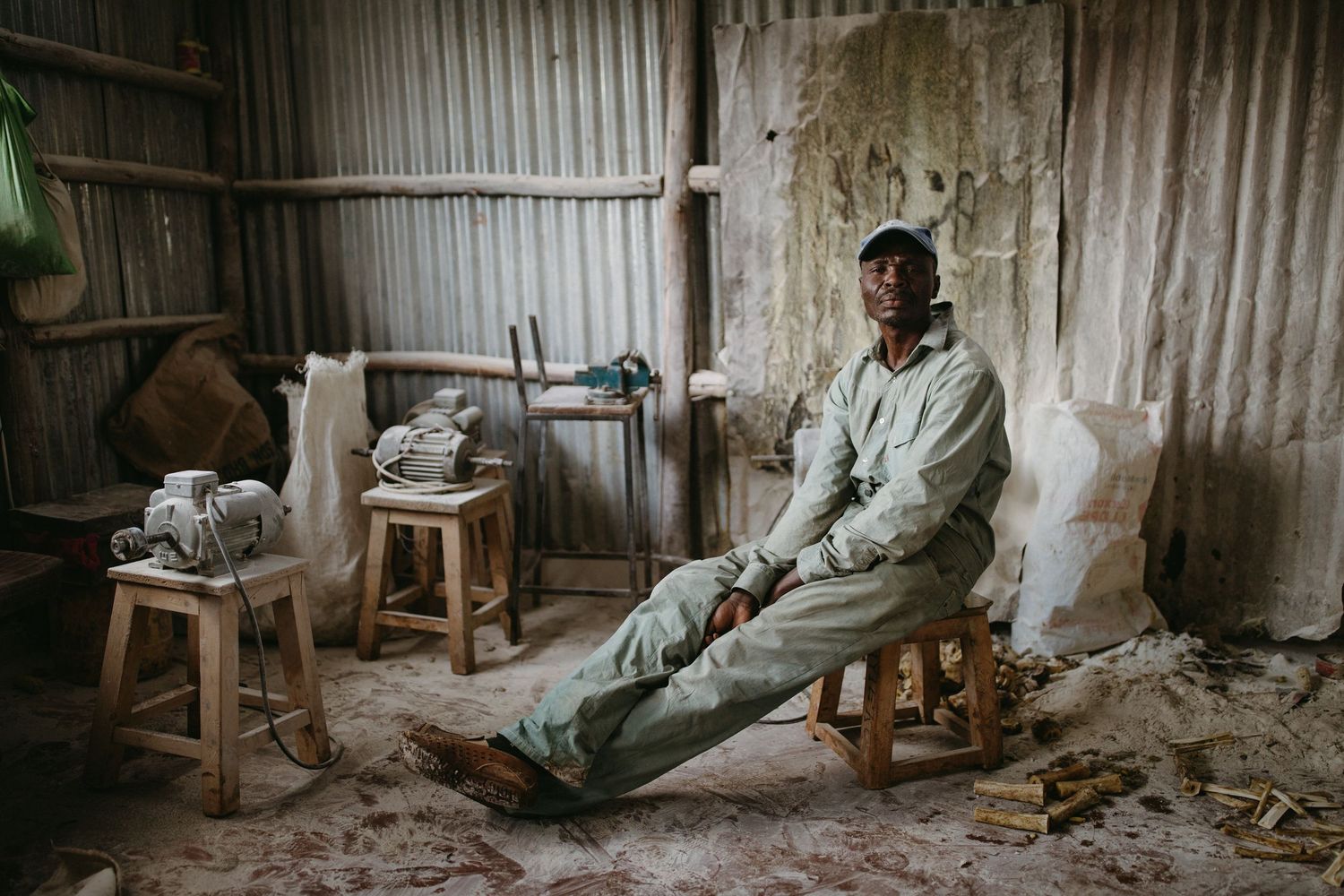 Figure seated on small wooden stool in rustic workshop with metal walls and scattered tools and materials.