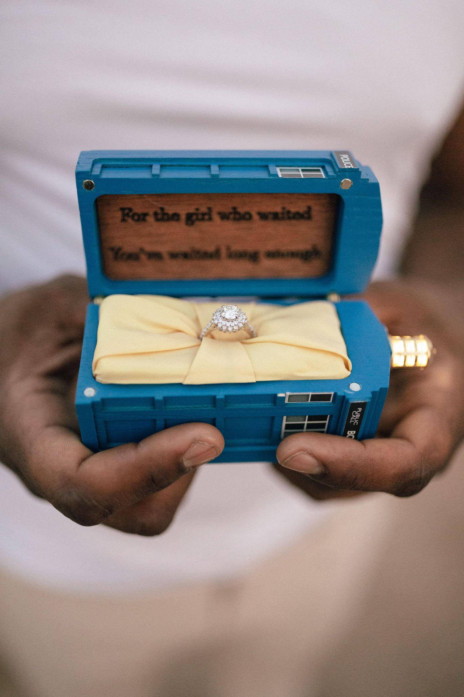Hands holding a blue TARDIS-style ring box with a diamond engagement ring nestled on yellow fabric inside.