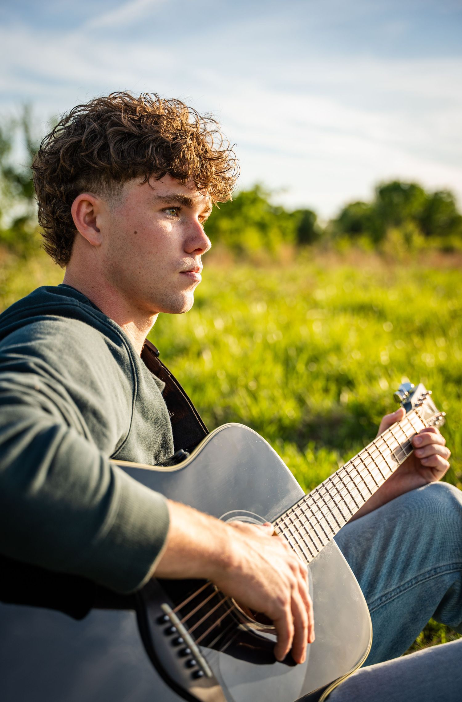 Person playing acoustic guitar outdoors in golden sunlight wearing casual grey attire.