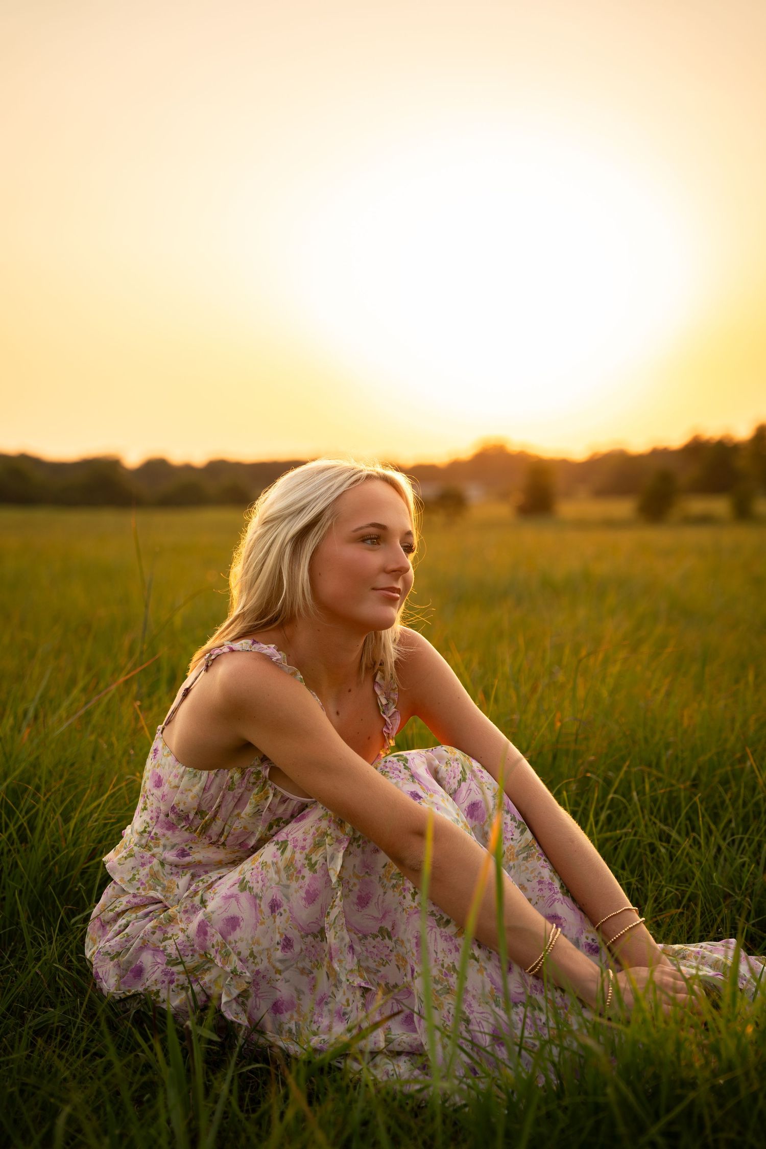 A person sits in a grassy field at sunset wearing a floral dress while enjoying the golden hour atmosphere.