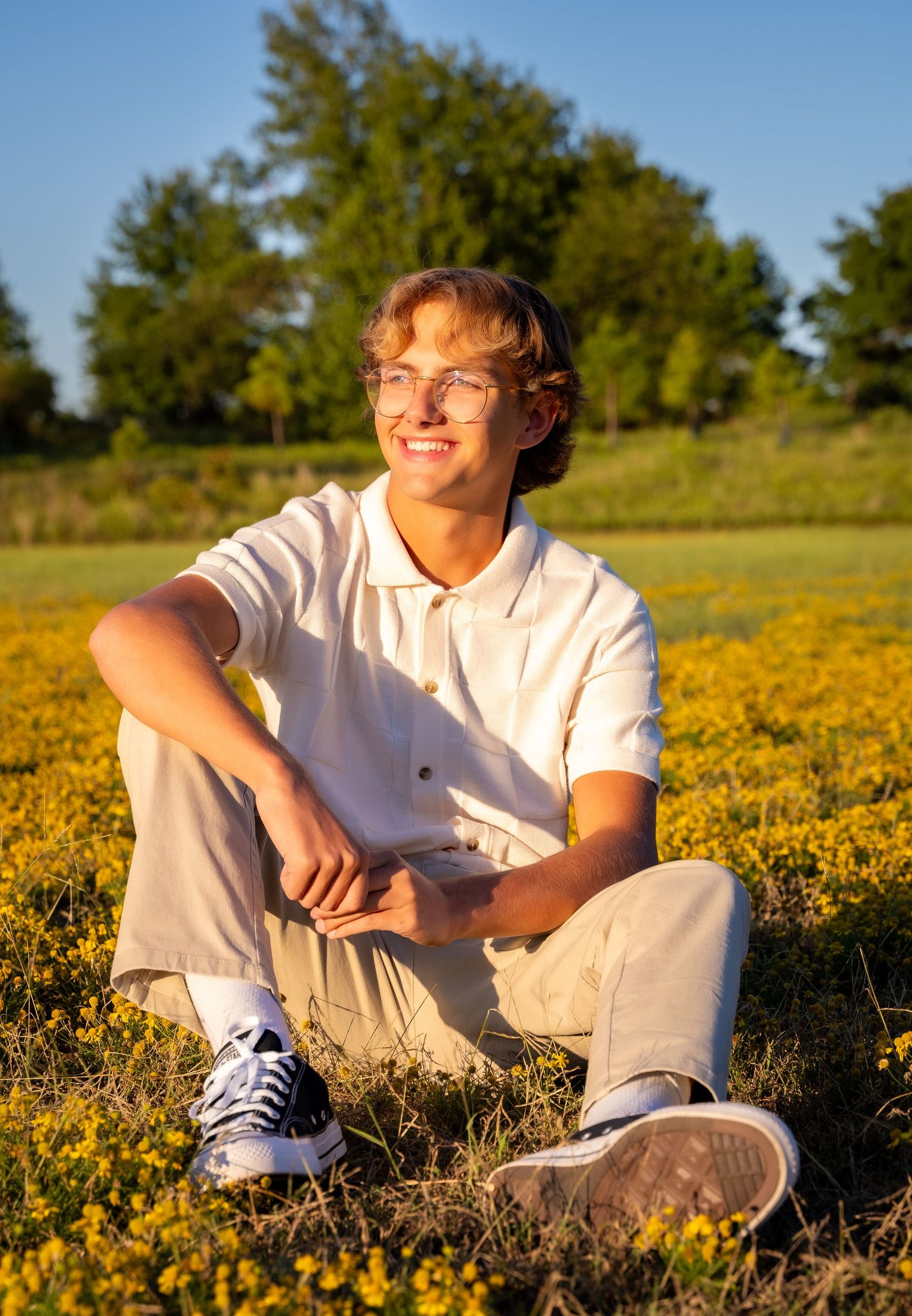 Person sitting in a field of yellow wildflowers at golden hour wearing glasses and casual attire.