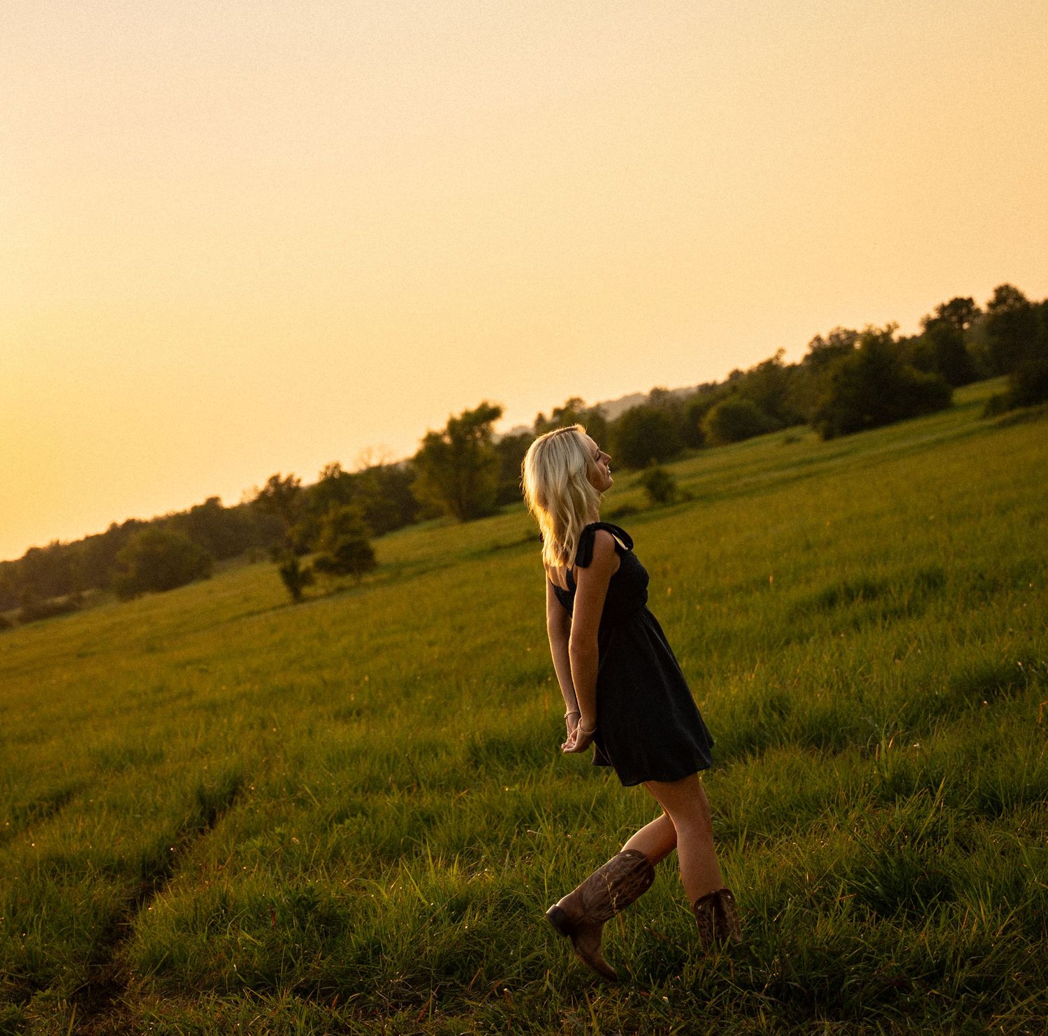 A silhouette walks through a grassy hillside field at golden hour with a warm sunset sky in the background.