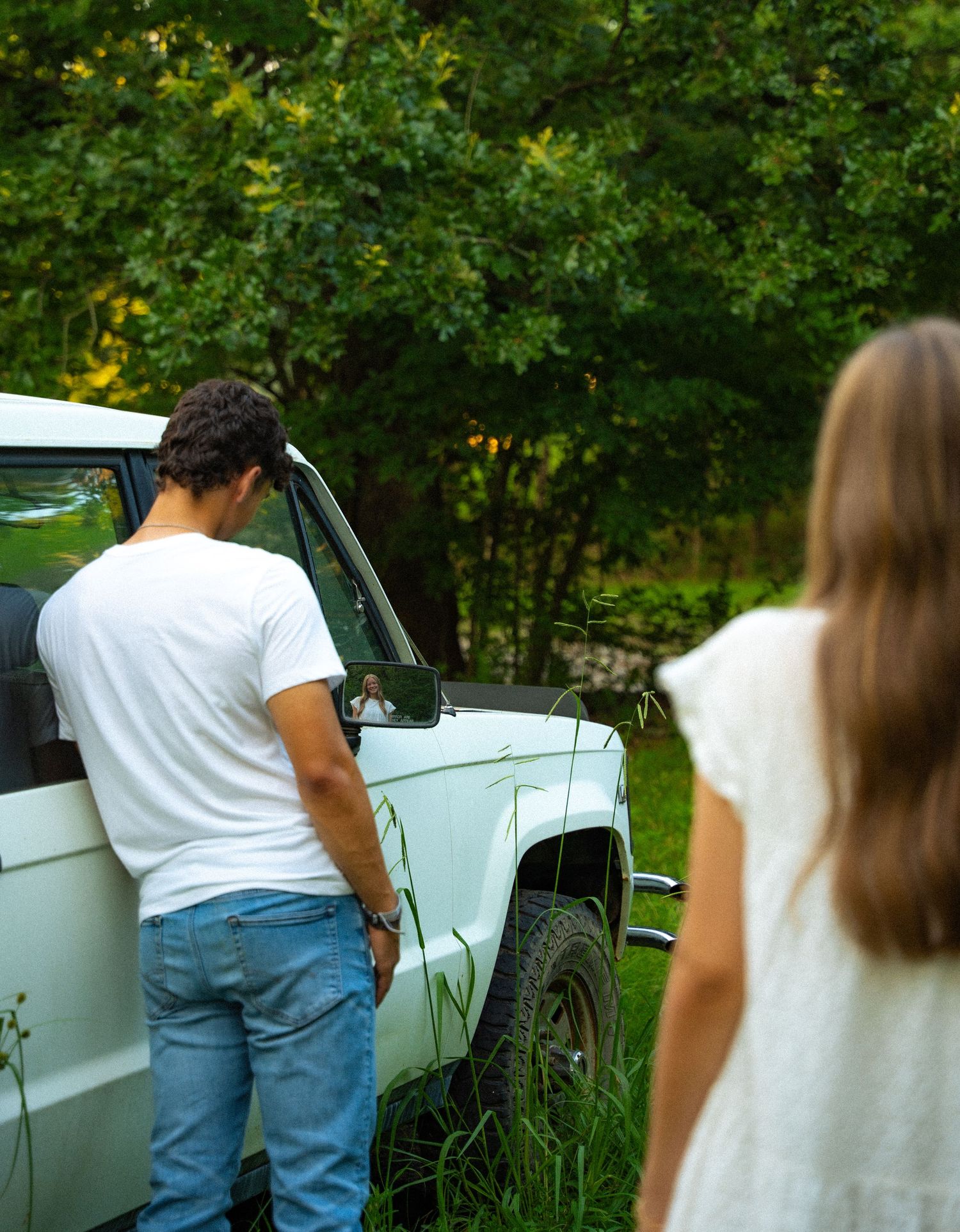 People stand by an old white vintage car parked in a lush green garden setting.