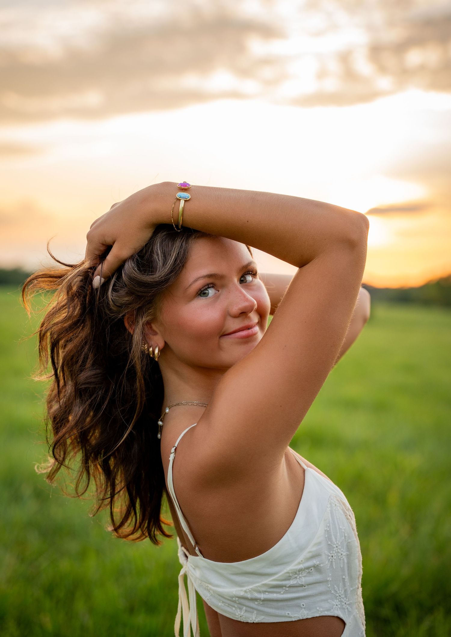Person with flowing hair poses gracefully against a golden sunset in a grassy field wearing a white sleeveless top.