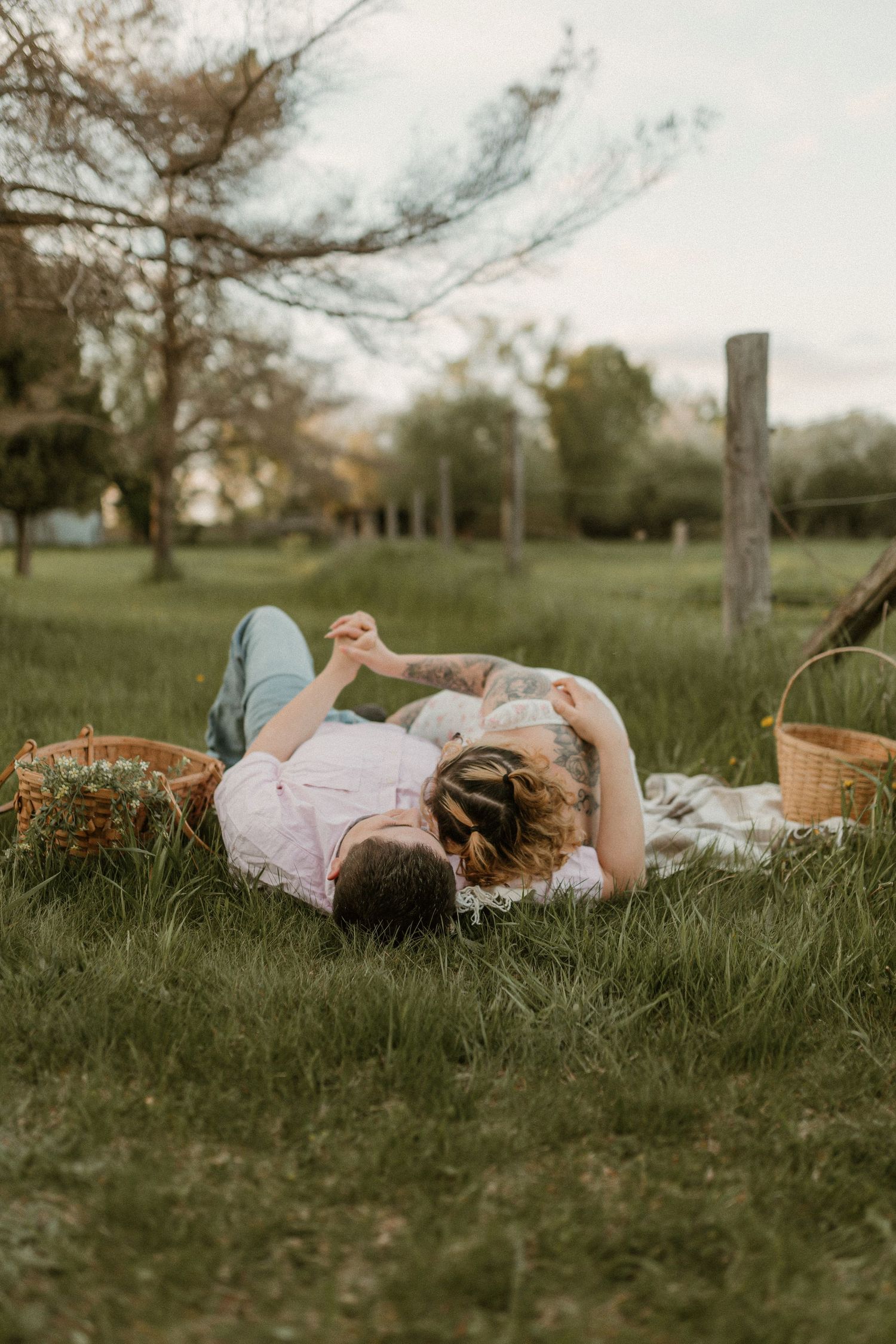 A couple lying on a blanket in grass during sunset with picnic baskets nearby.