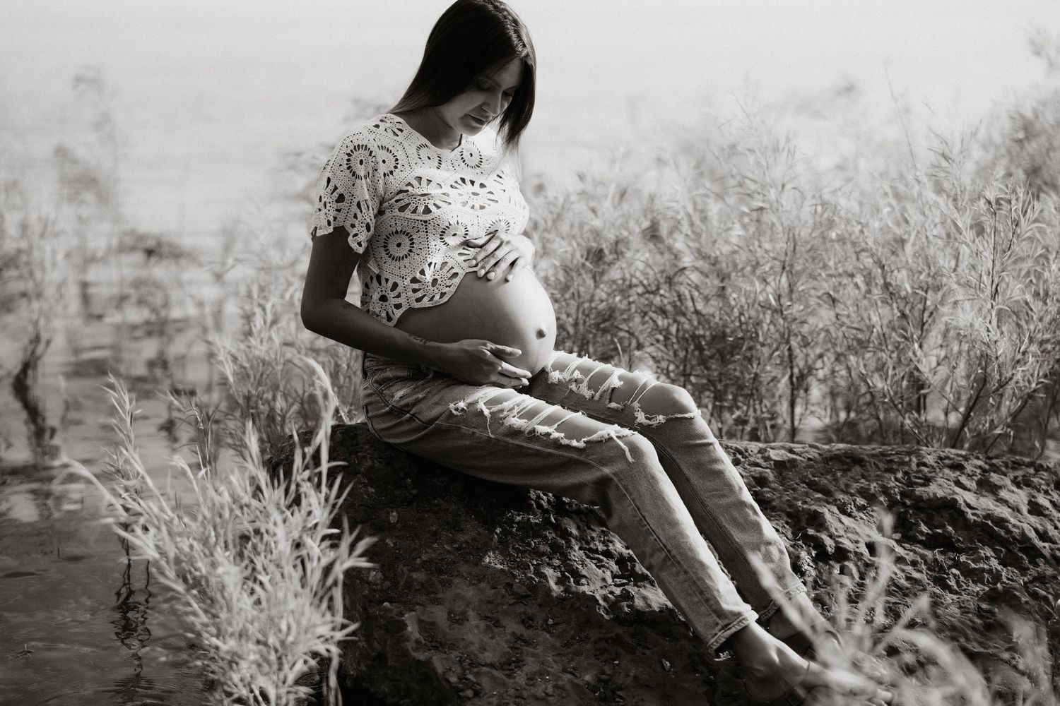 Artistic black and white maternity photo of woman sitting by water.