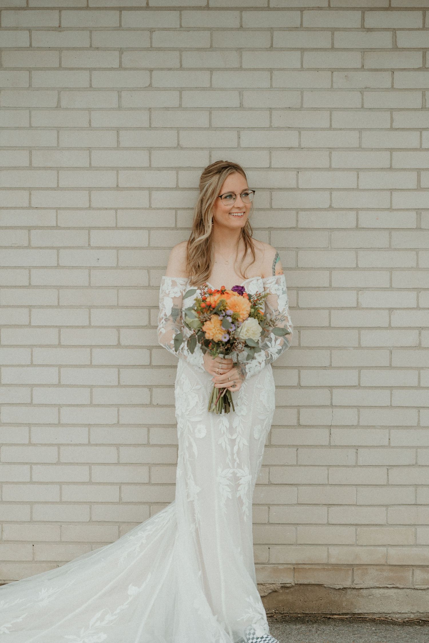 Bride in lace white gown holds colorful bouquet while posing against white brick wall.