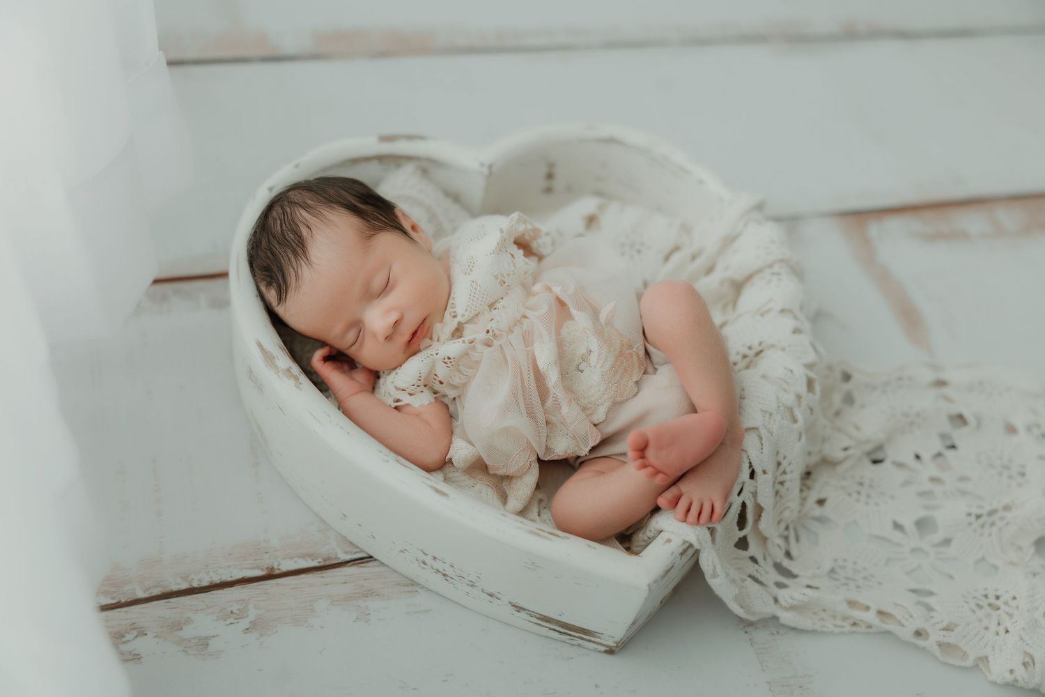 Sleeping baby in lace outfit nestled in heart-shaped white prop.