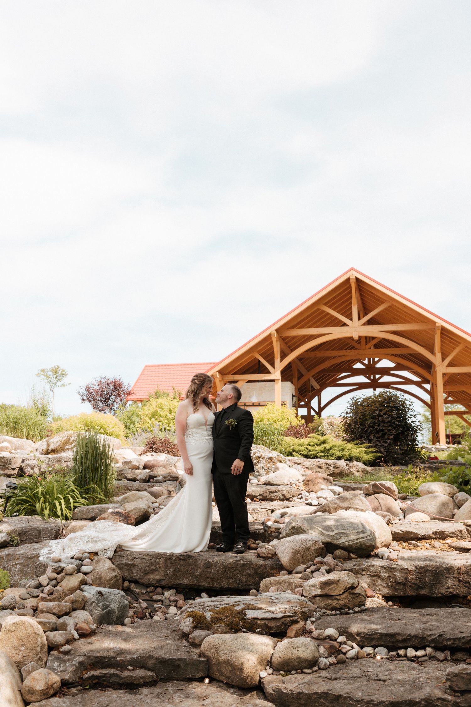 A wedding couple poses on rocky landscape with wooden pavilion in background.