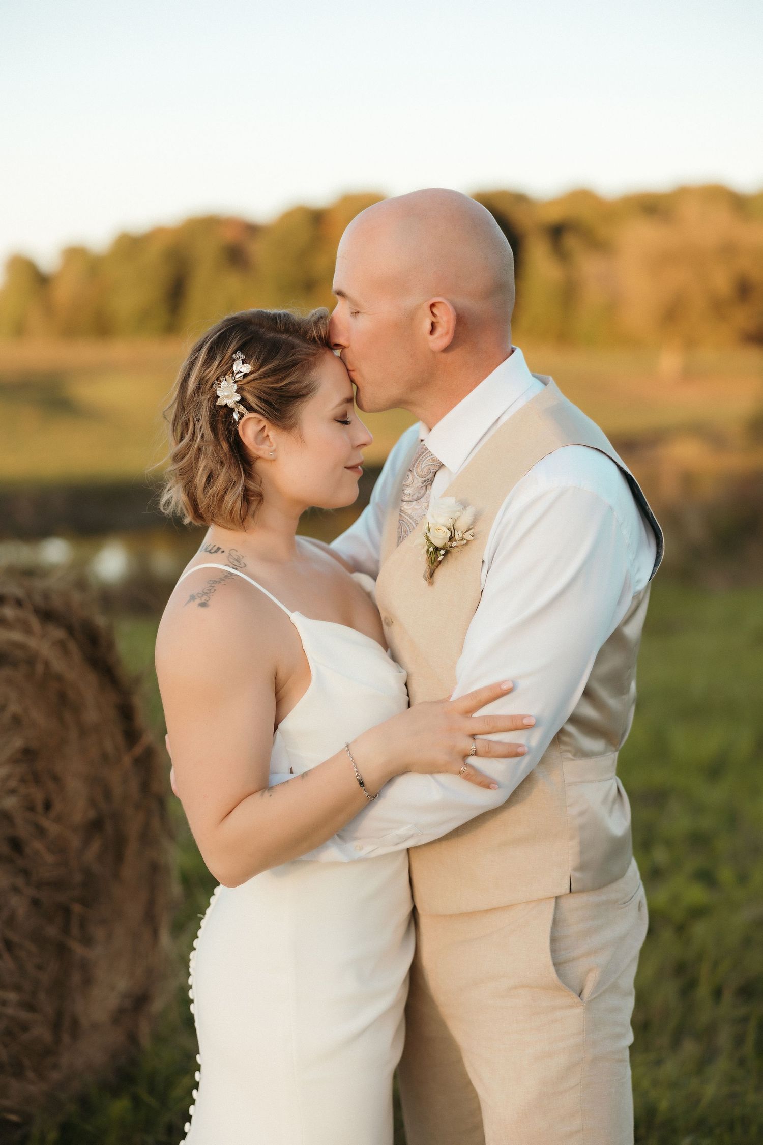 Couple in wedding attire embracing in an outdoor field at sunset.