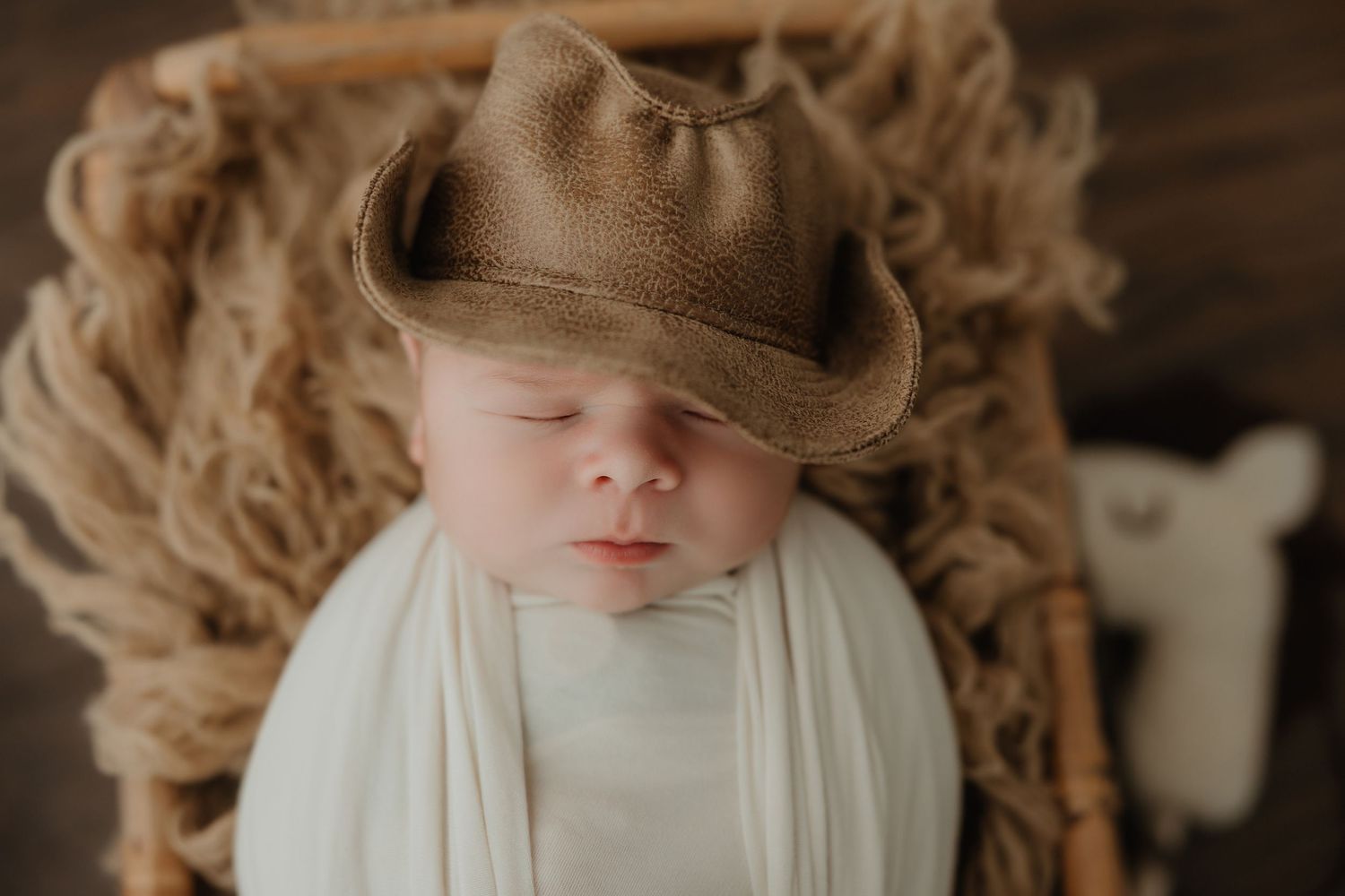 Sleeping newborn wearing brown cowboy hat on textured blanket.