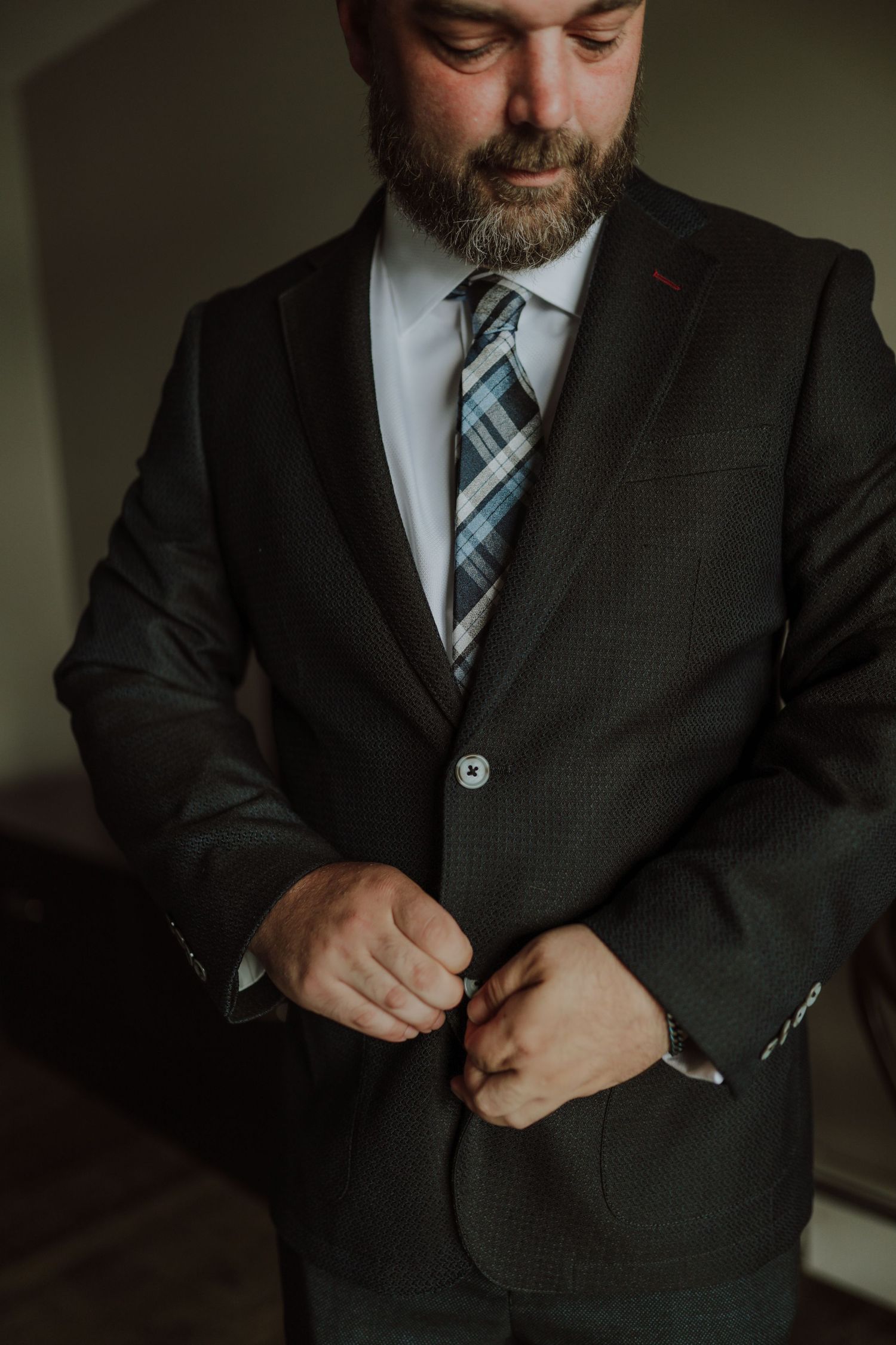 A groom in a dark suit and plaid tie adjusts his jacket buttons while getting ready.