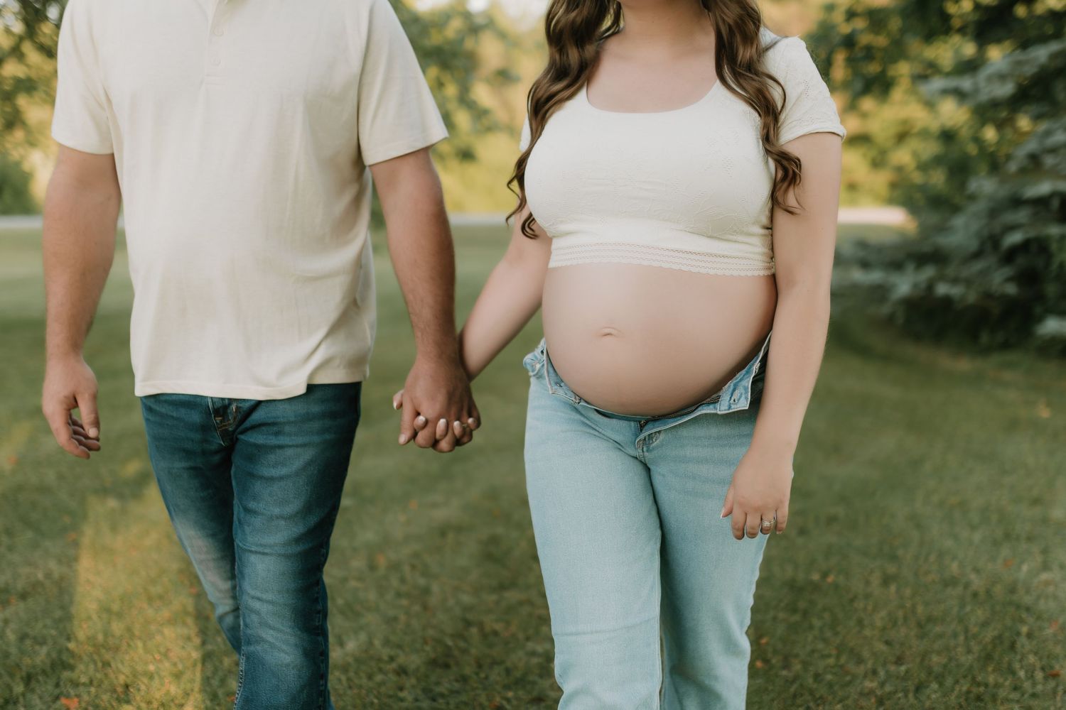 A couple holds hands during their maternity photoshoot in a lush green outdoor setting.