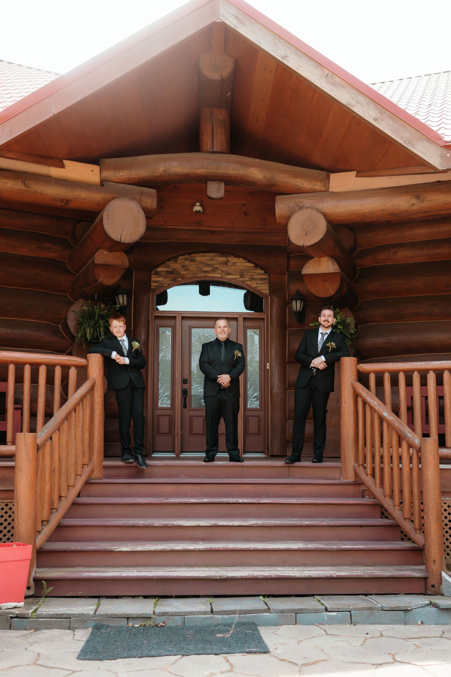 Three groomsmen stand on the porch of a rustic log cabin entrance.