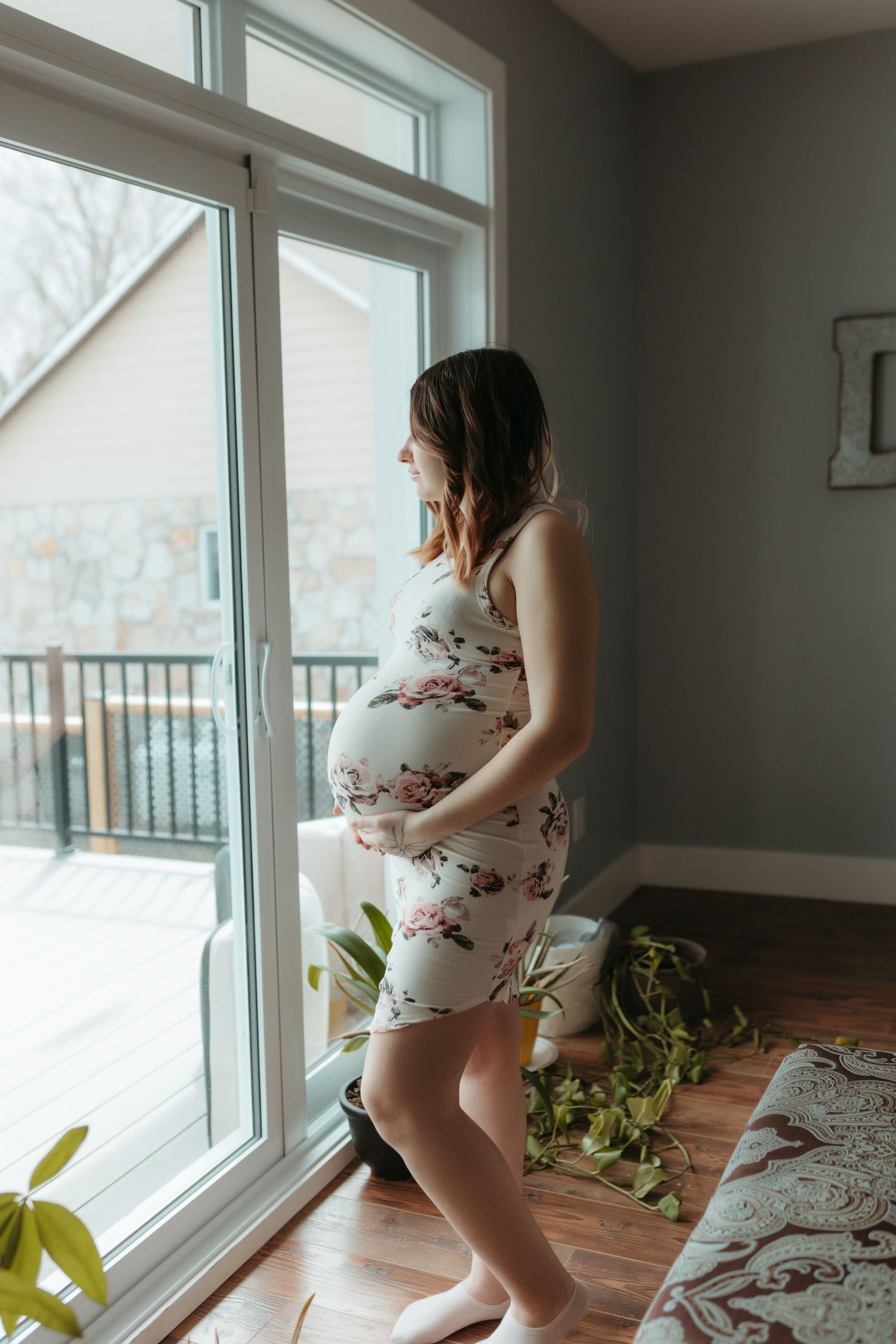 Maternity photos near sunlit windows with greenery and a floral dress creating a bright, natural atmosphere.