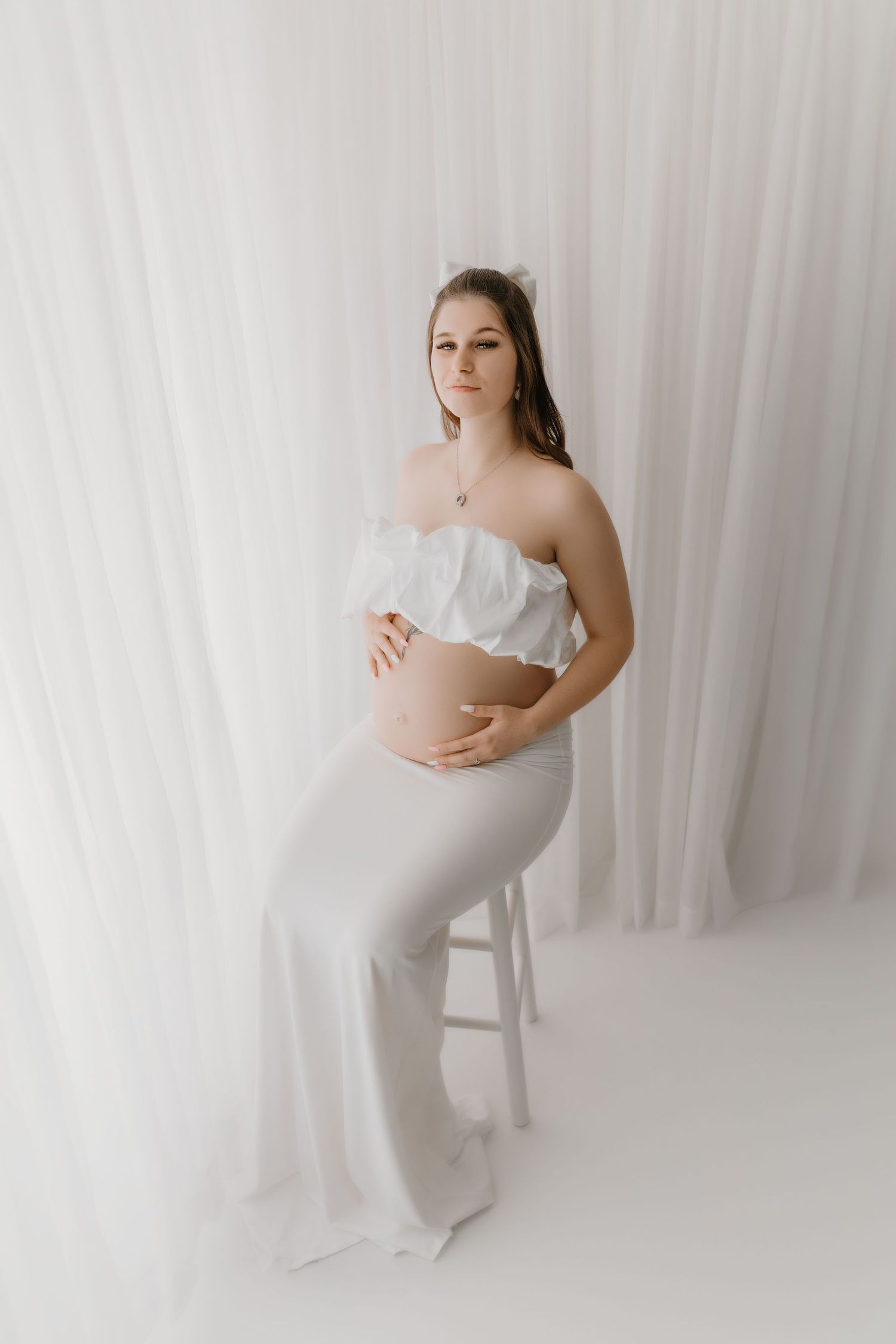 Maternity portrait of woman in white outfit against light backdrop.
