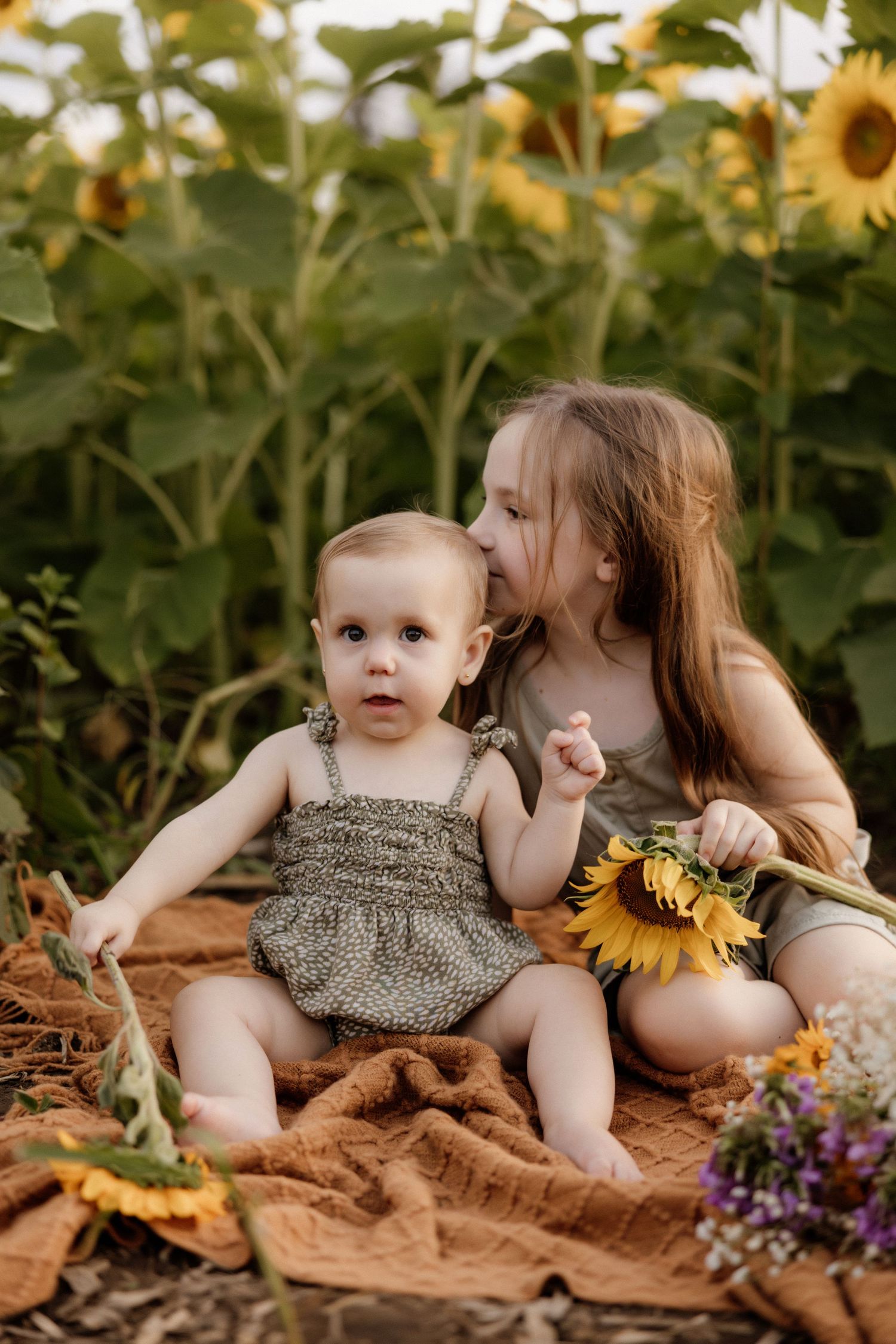 Two children sitting on a blanket among sunflowers and wildflowers.