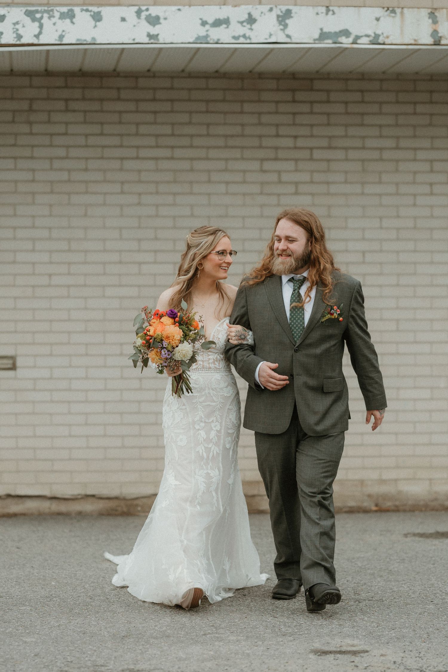 Newlyweds take candid photos walking together on a paved walkway beside white brick walls.