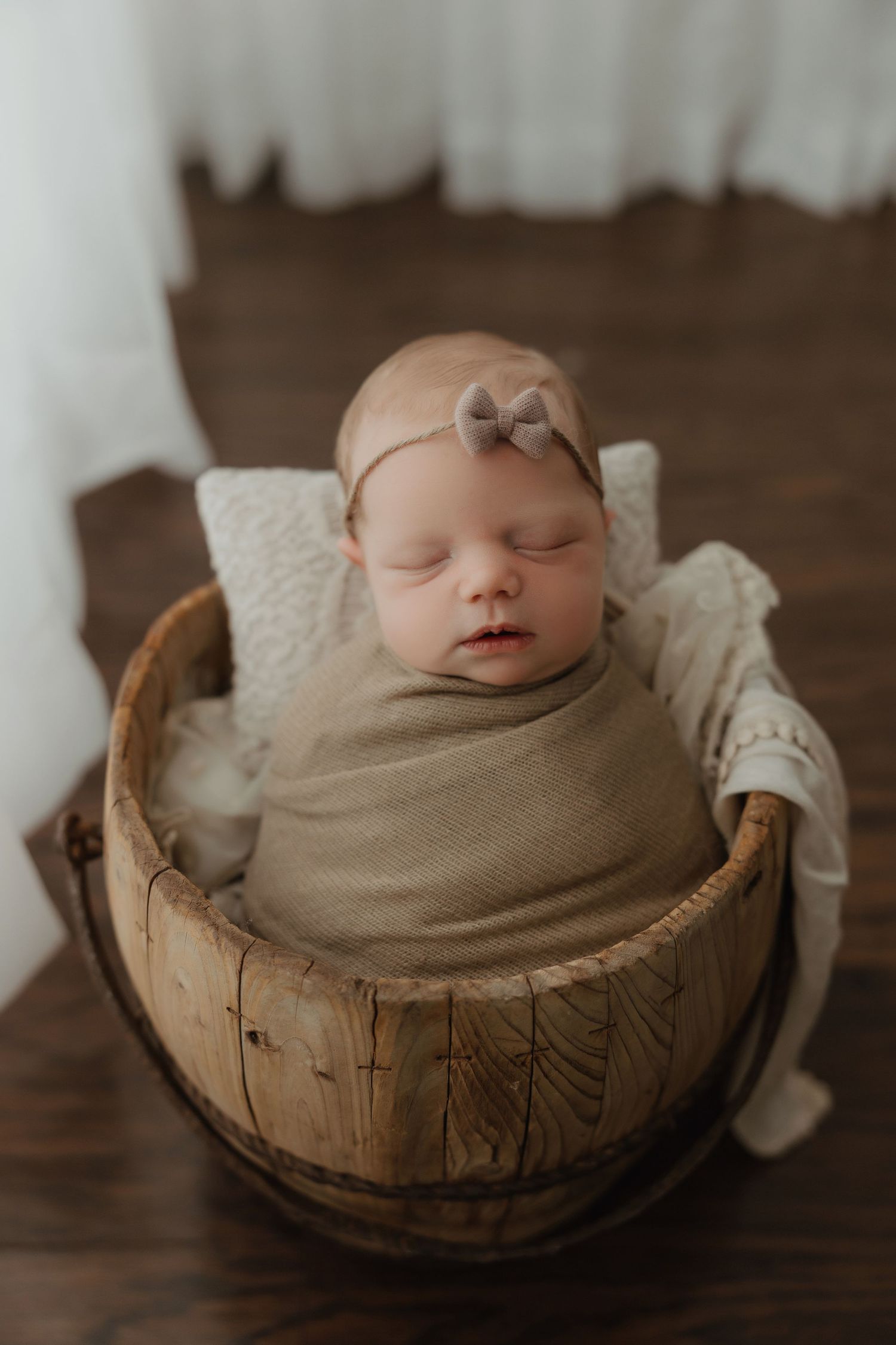 Peaceful newborn wrapped in neutral swaddle sleeping in wooden bowl on dark wood floor.