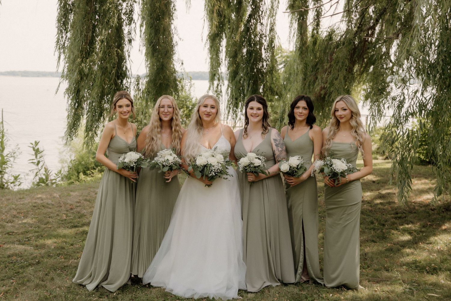 Bridal party in sage green dresses poses under weeping willow tree for wedding photos.