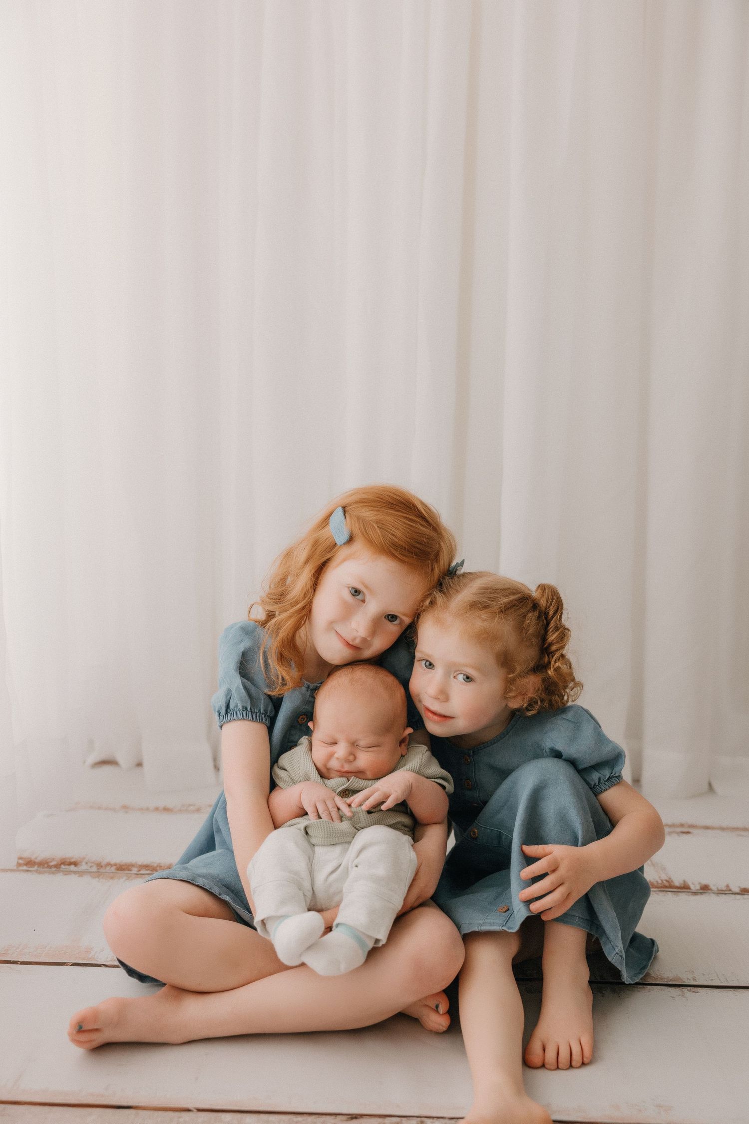 Two children in blue dresses holding newborn sibling against white backdrop.
