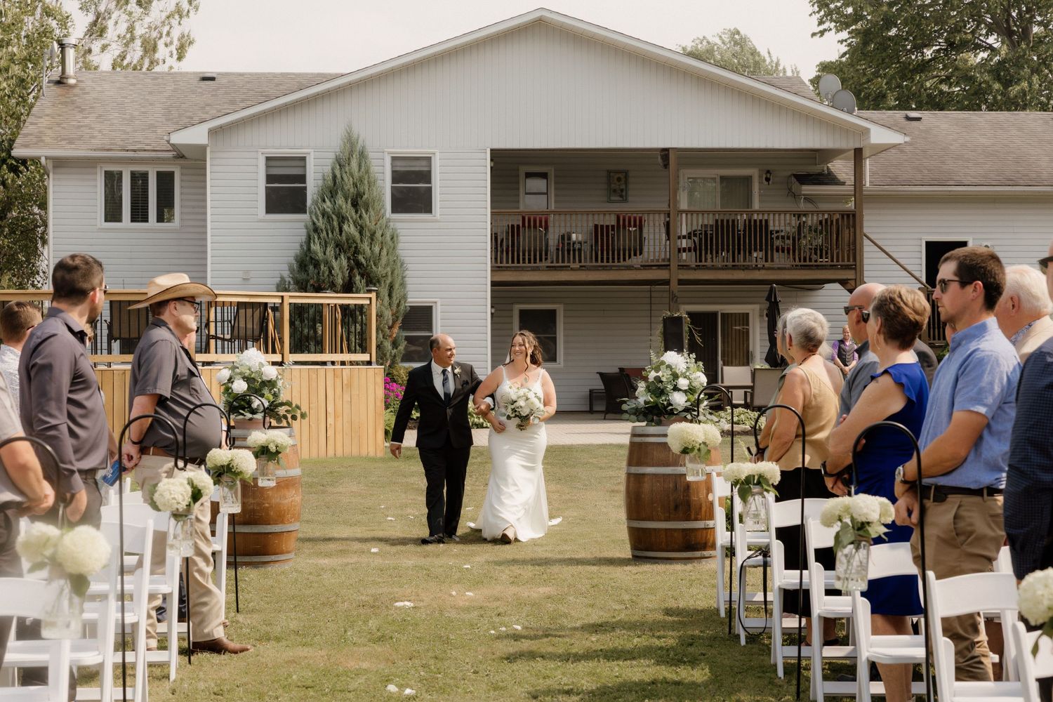 Outdoor backyard wedding ceremony with white chairs and guests watching processional.