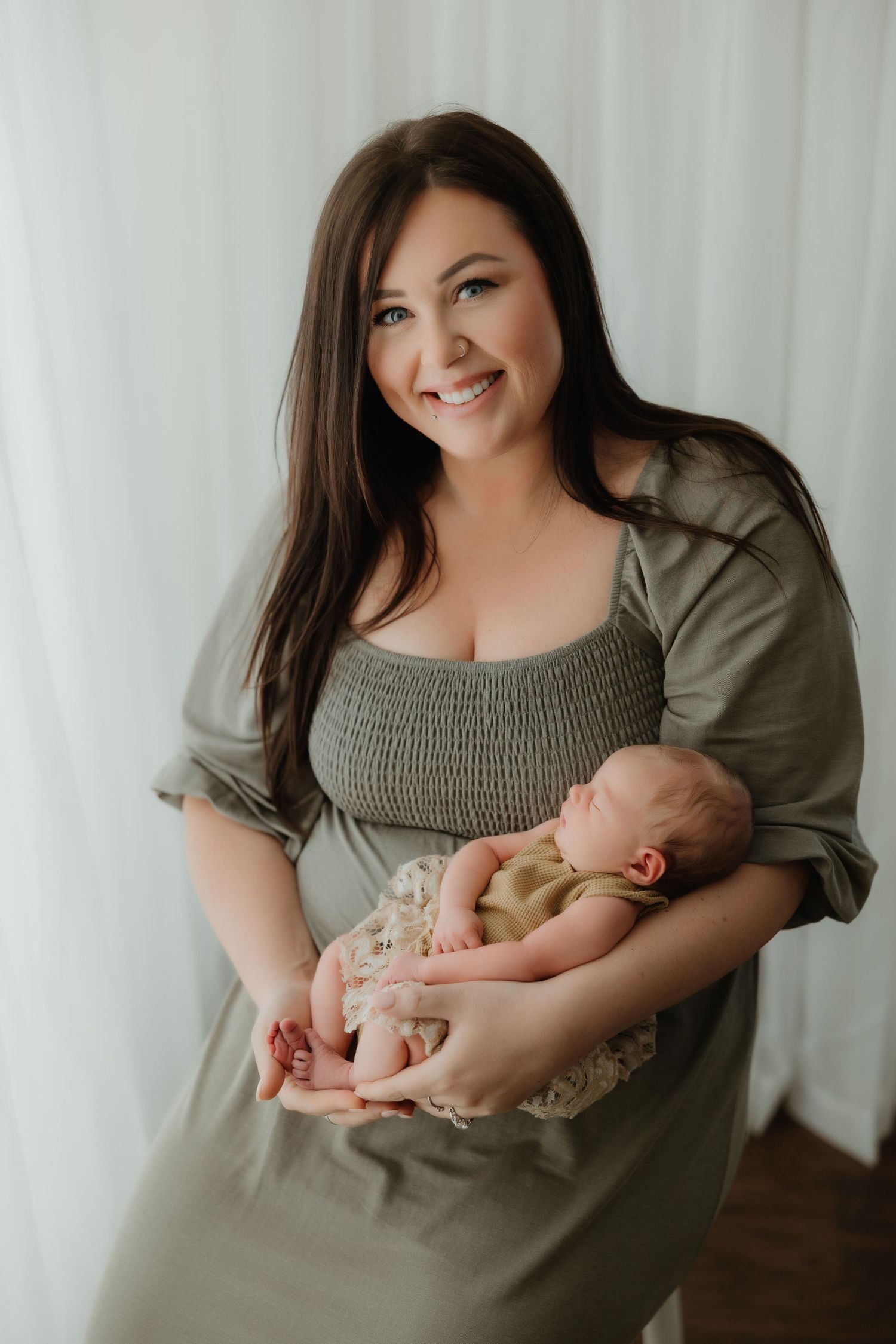 Woman in olive green dress holding a sleeping newborn baby against white curtain background.