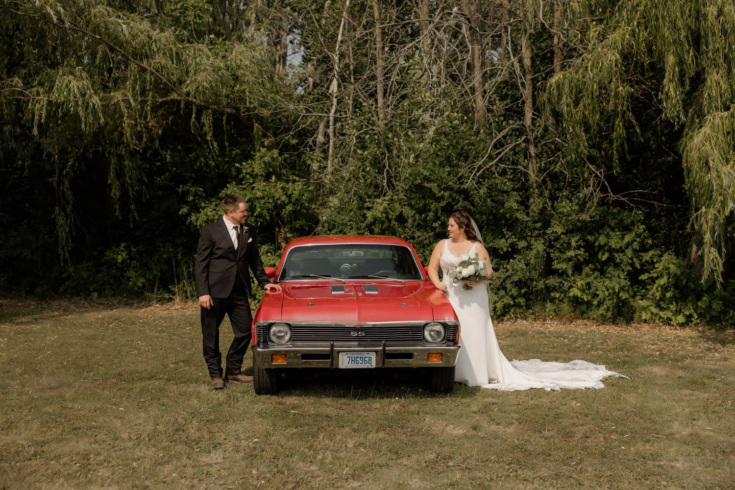 A classic red vintage car alongside a wedding couple in an outdoor garden setting.