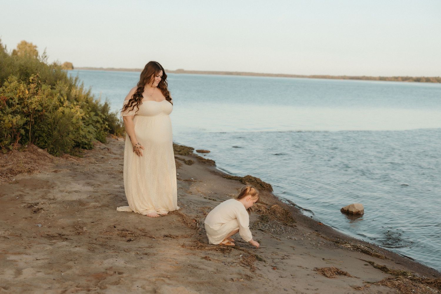 Expecting mother in white dress stands by water while child plays on shore.