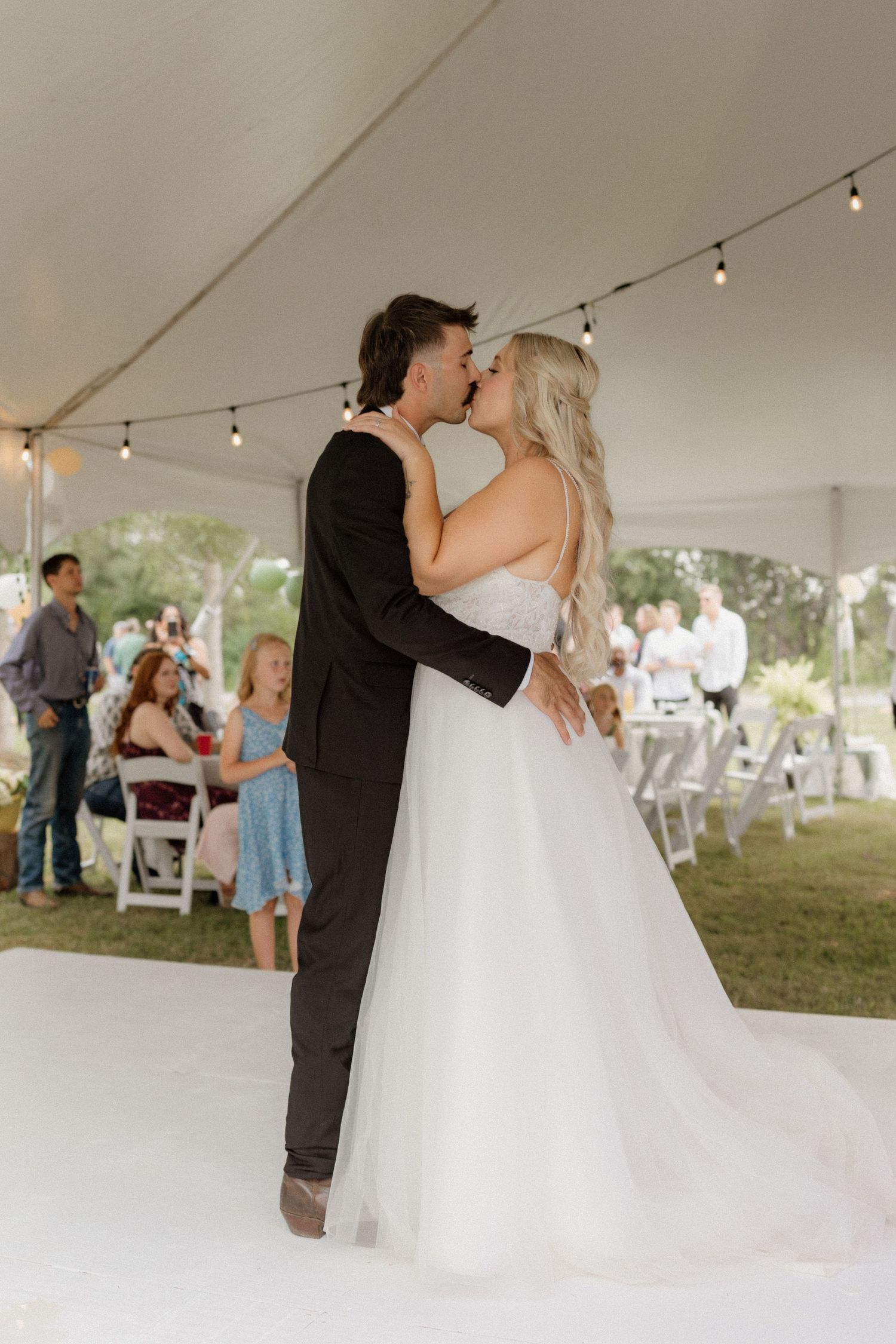 Wedding couple shares romantic first dance under string lights in outdoor tent reception.