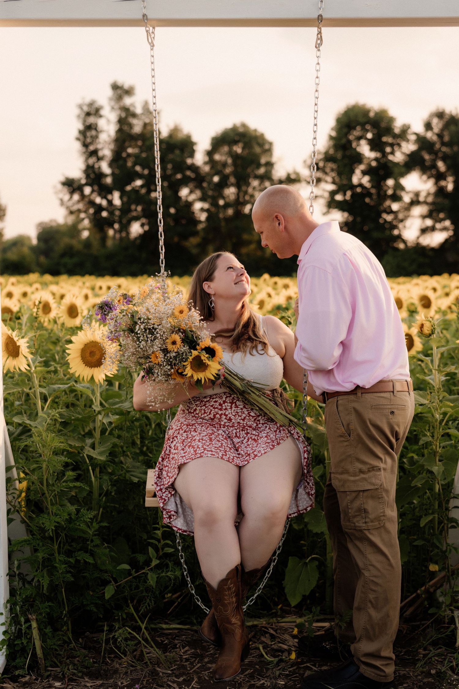 Romantic couple poses on a swing in a sunflower field during golden hour.
