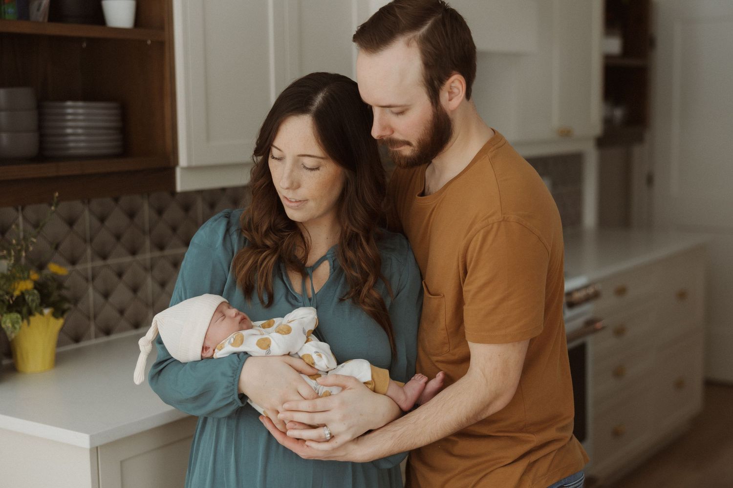 Parents cradle their newborn baby in a cozy kitchen setting with natural lighting.