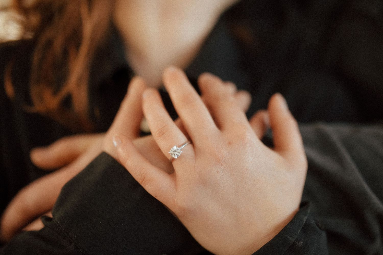 A hand wearing a sparkly diamond engagement ring against a dark background.