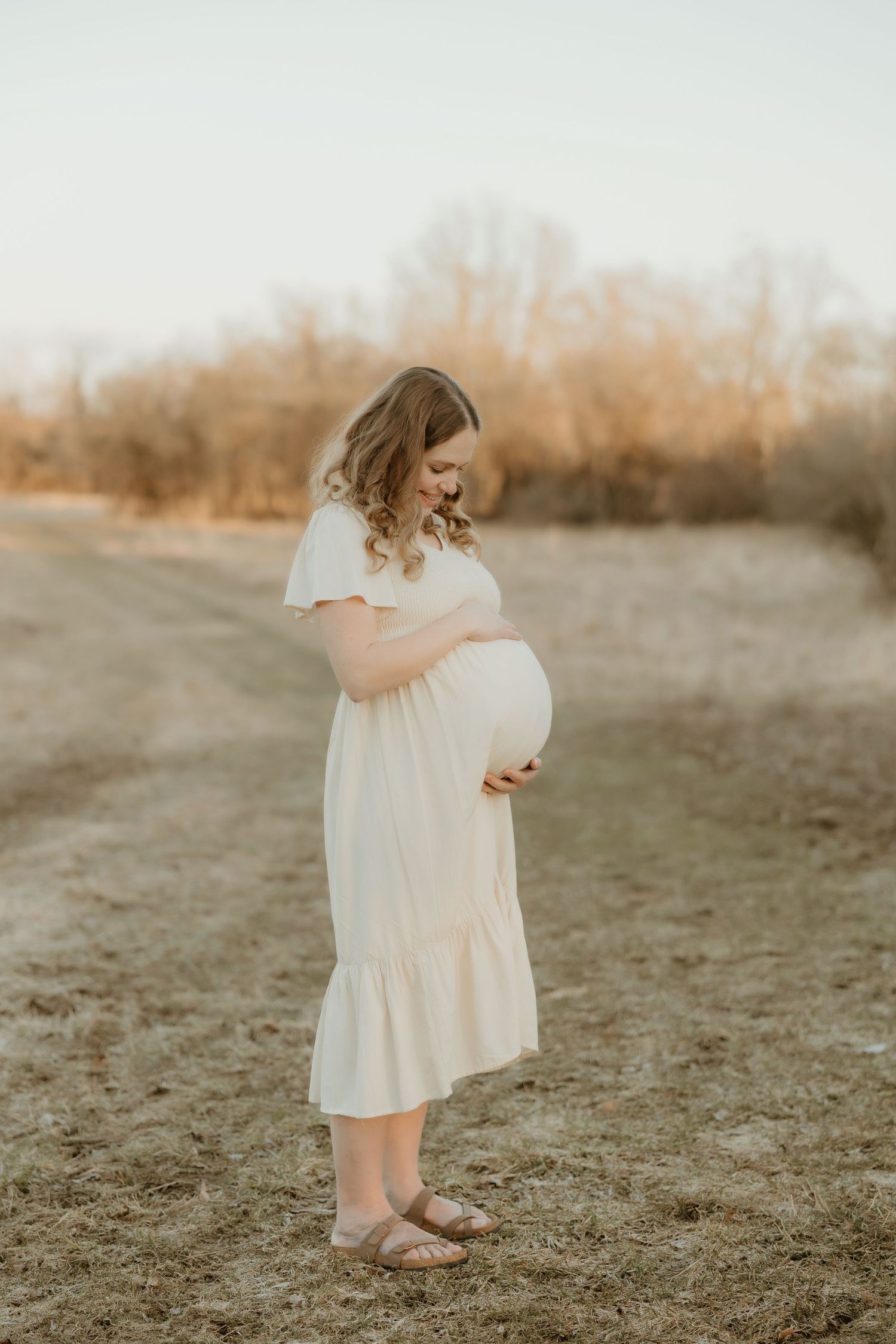 Woman in flowing white dress cradles baby bump during golden hour maternity photo shoot.