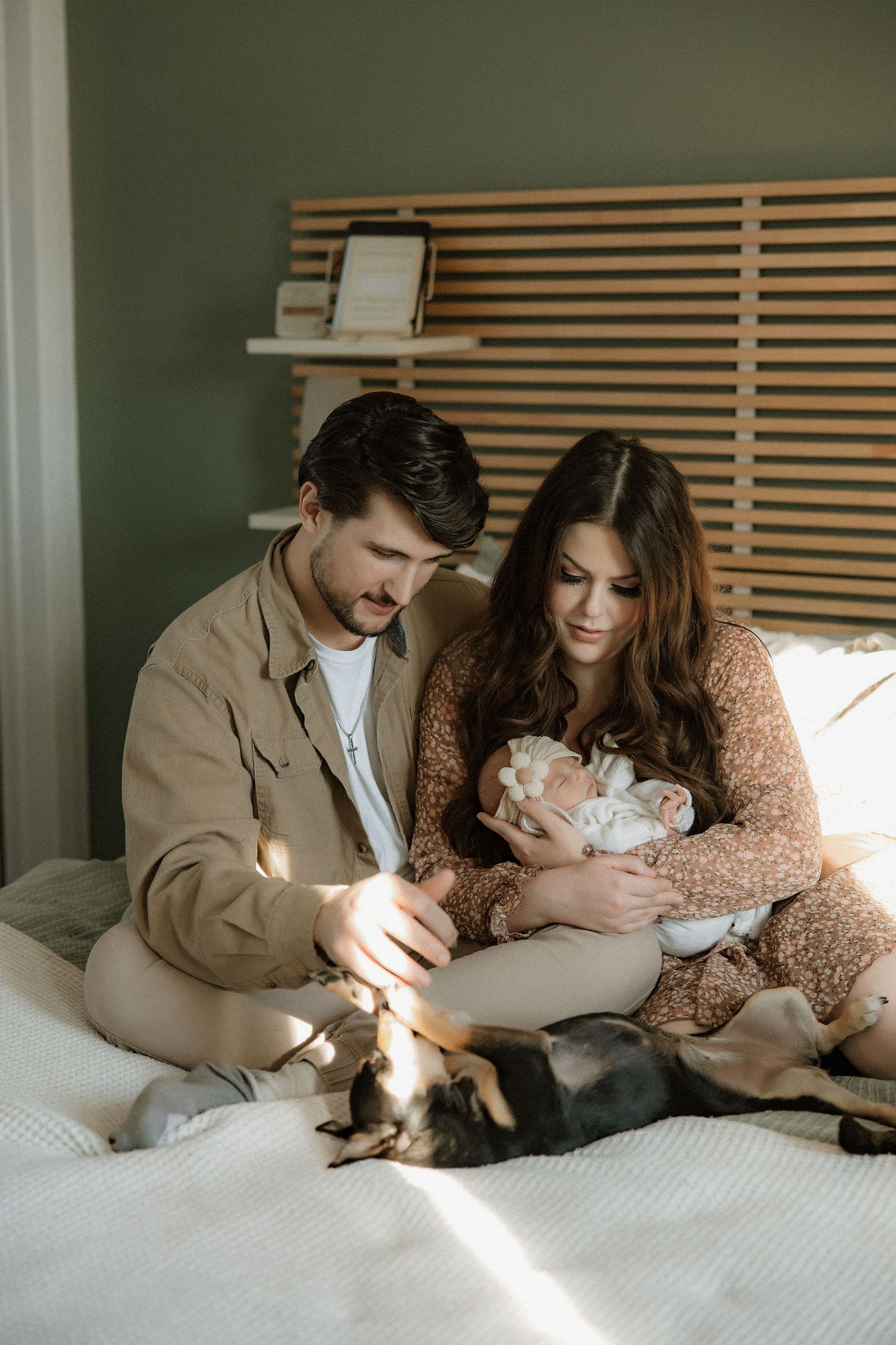Family lounging together on white bedding with sunlight filtering through wooden blinds.