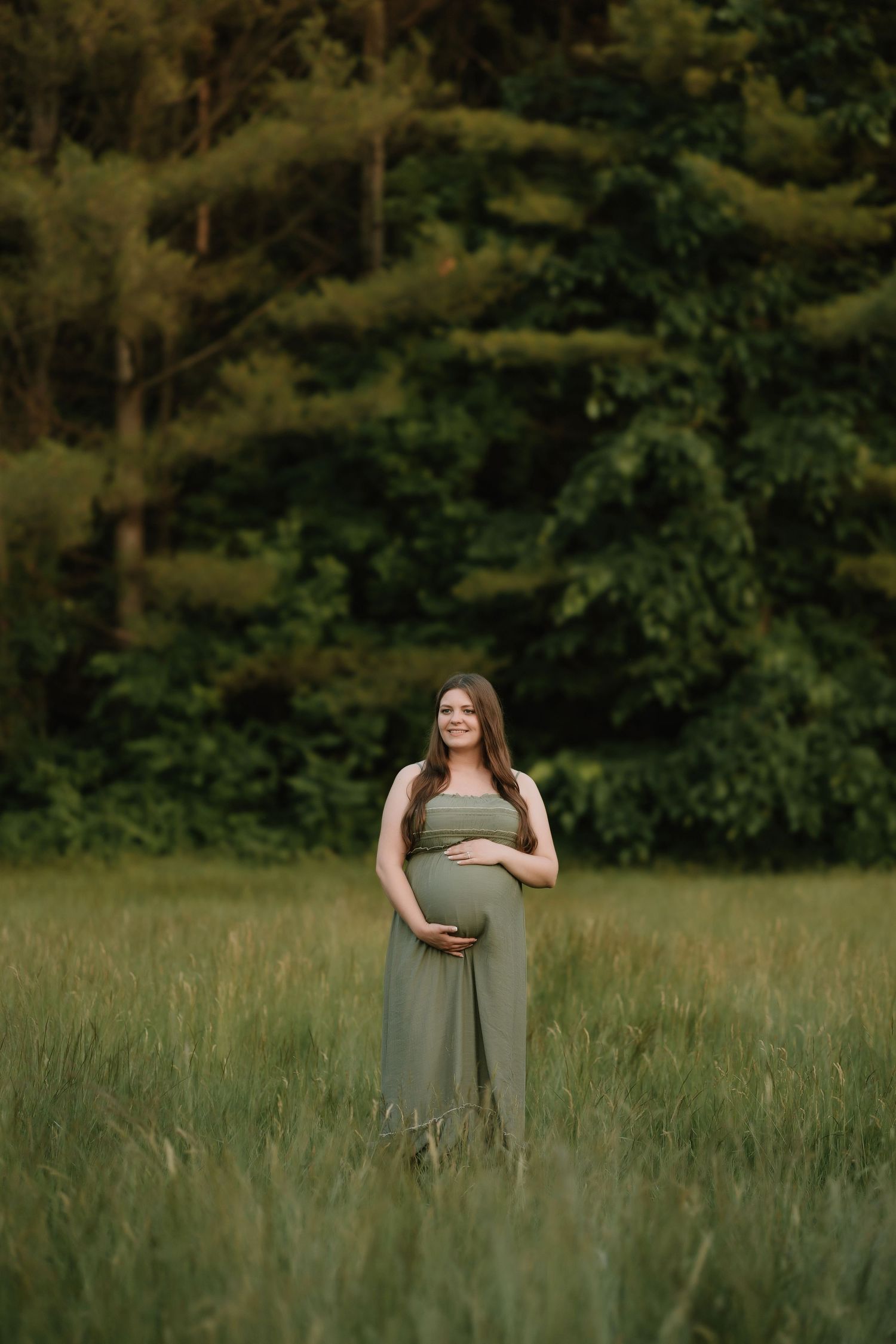 Expectant mother in olive dress standing in tall grass during sunset photoshoot.