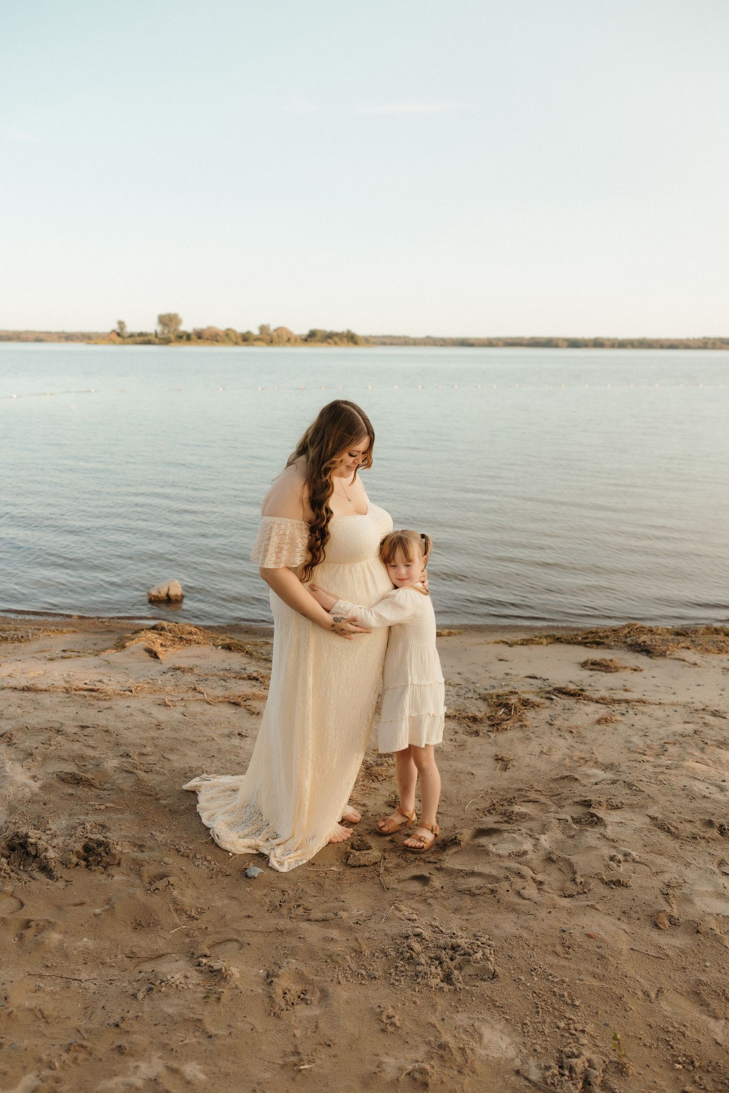 A mother and child stand on a sandy beach at sunset wearing flowing white dresses by a serene lake.