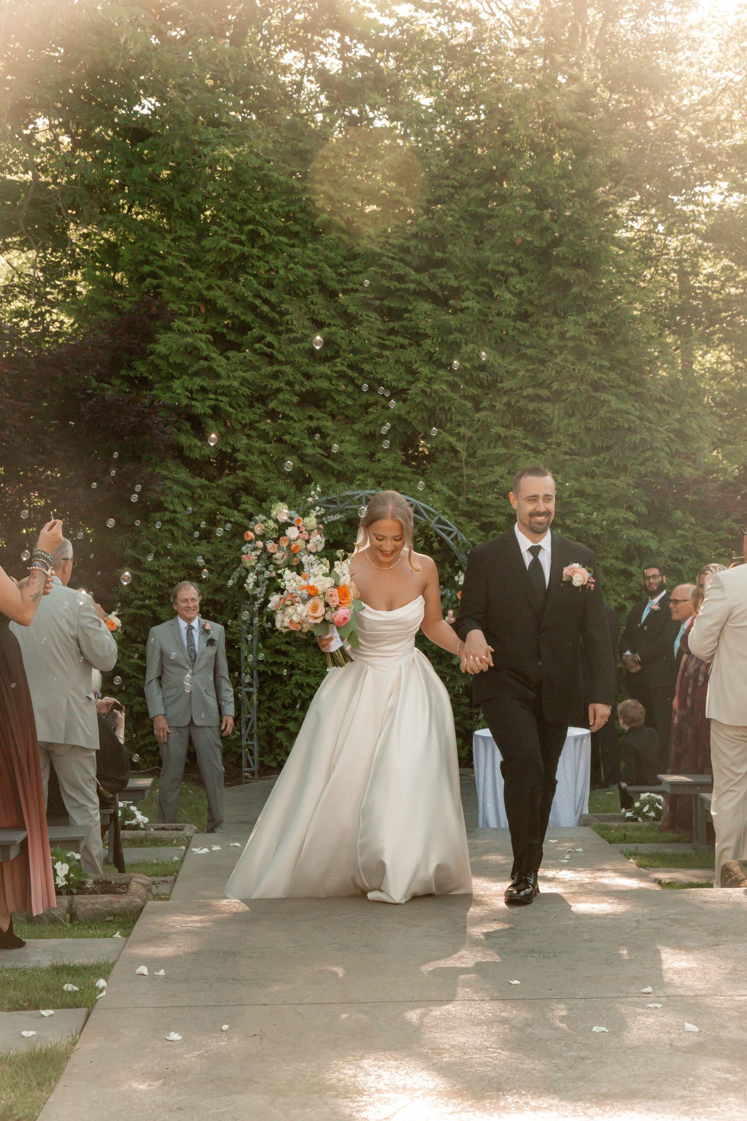 Bride and groom walk down outdoor aisle after wedding ceremony, surrounded by guests, lush greenery, and golden sunlight.