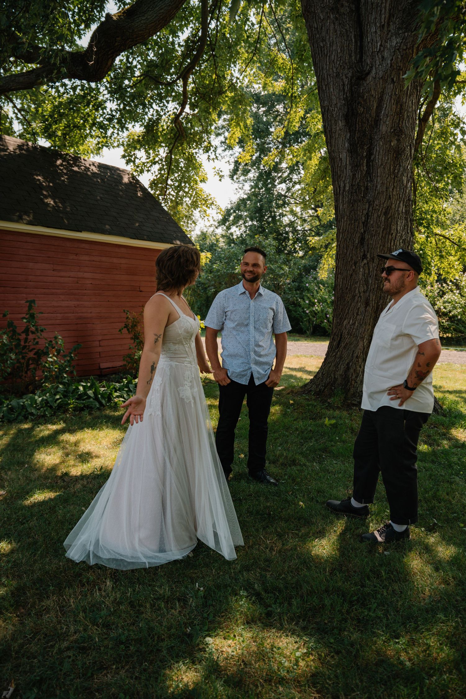 Bride in white gown talks with two men under large tree near red barn during outdoor wedding celebration.
