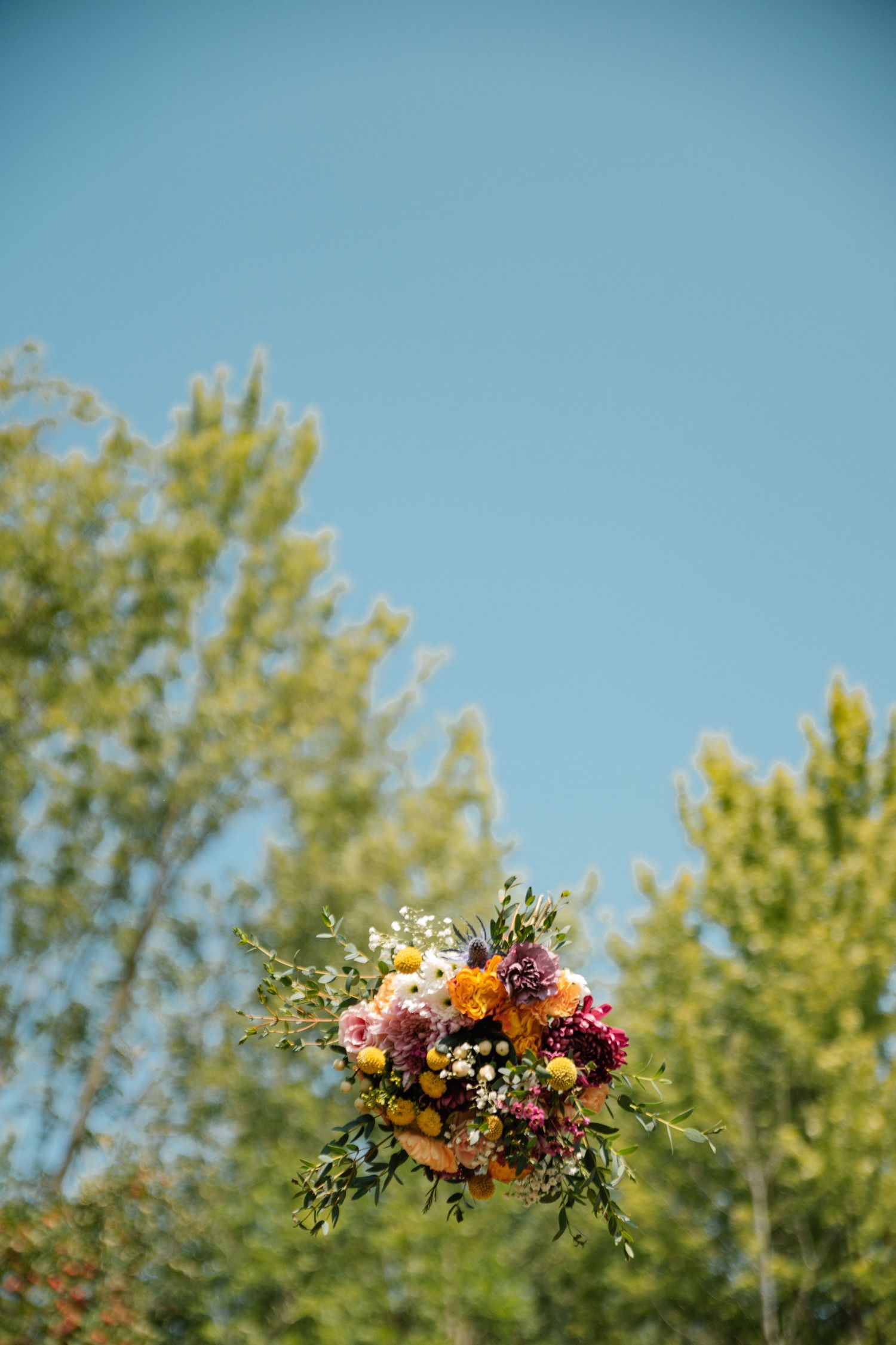 Colorful wedding bouquet with yellow, pink, and white flowers suspended against a clear blue sky with green trees.