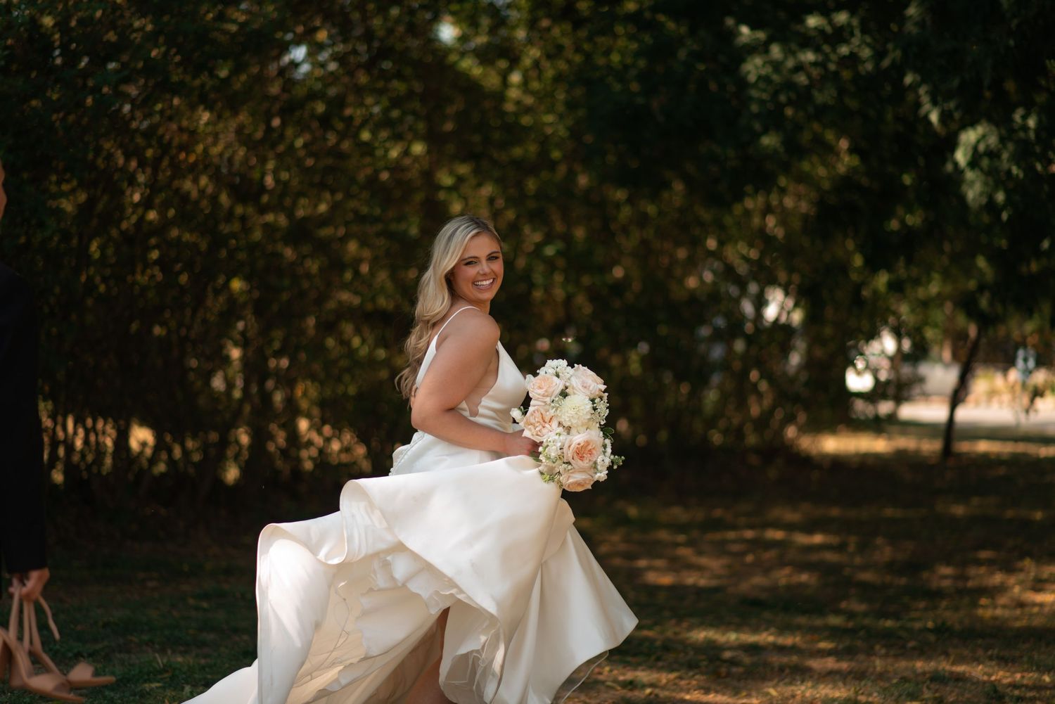 Smiling bride in white gown holding bouquet of white flowers outdoors among lush green trees.