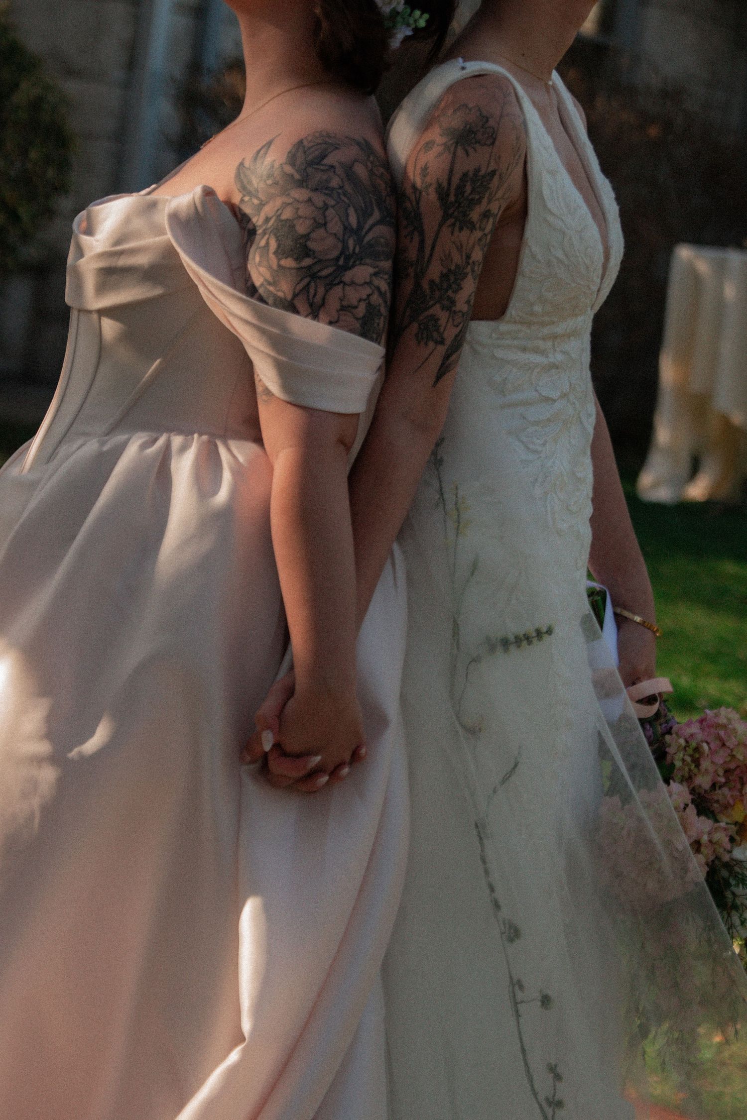 Two brides in white wedding gowns with floral tattoos hold hands back-to-back in an outdoor ceremony setting.