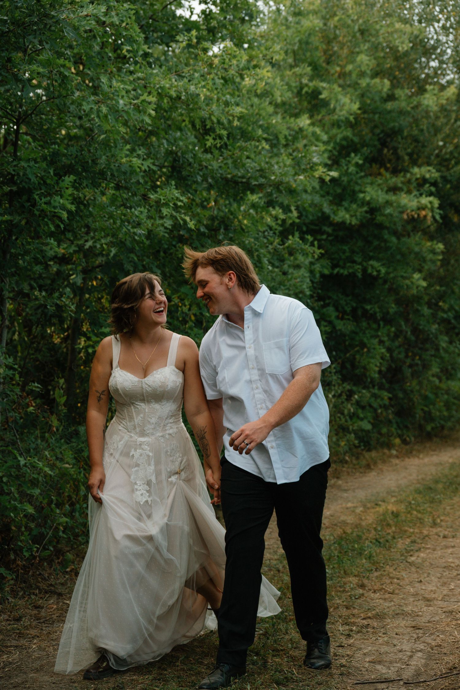 A laughing bride in a lace gown and groom in white shirt walk hand in hand on a wooded path.
