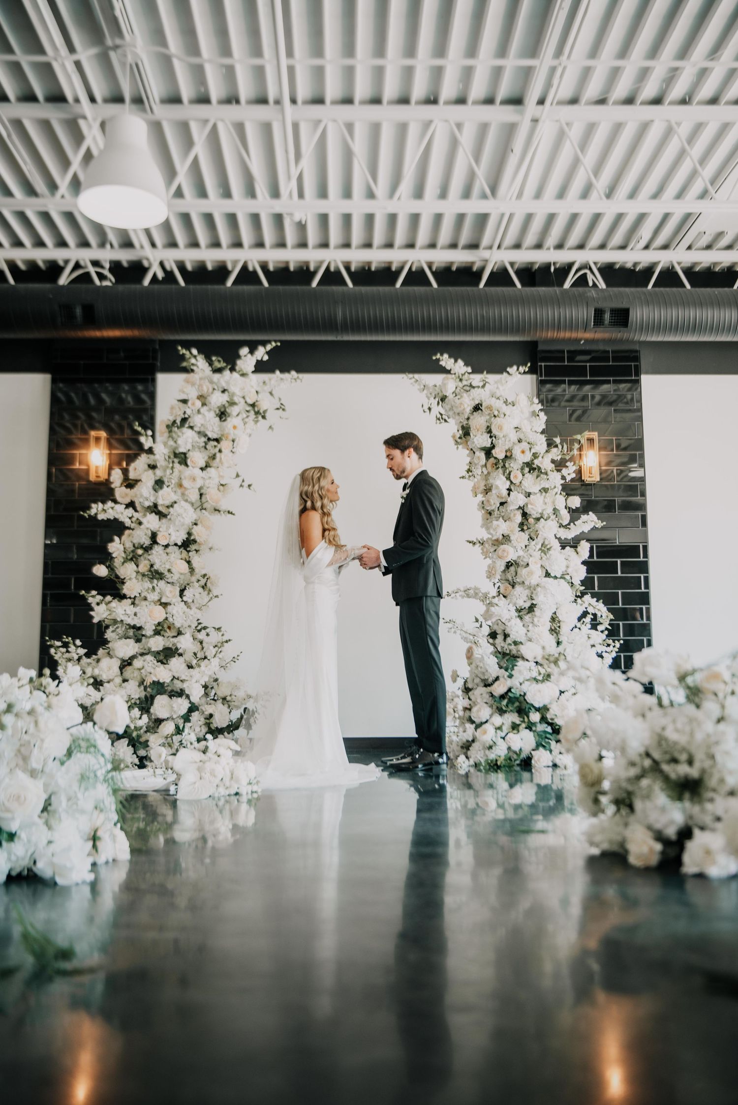 A modern industrial wedding ceremony space with white floral arch installations and black brick walls.