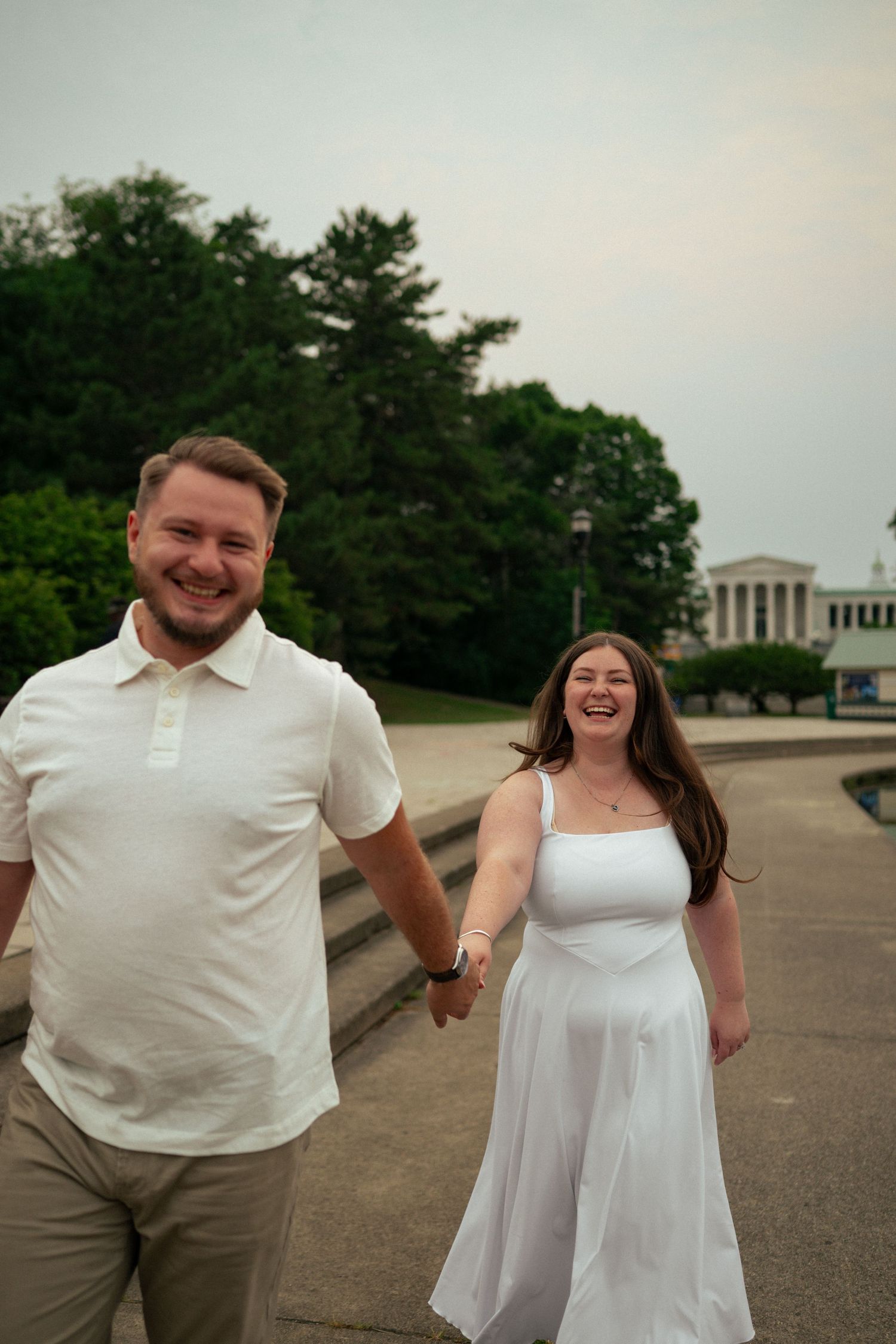 A smiling couple holds hands and walks outdoors near a classical building, wearing white outfits on a cloudy day.