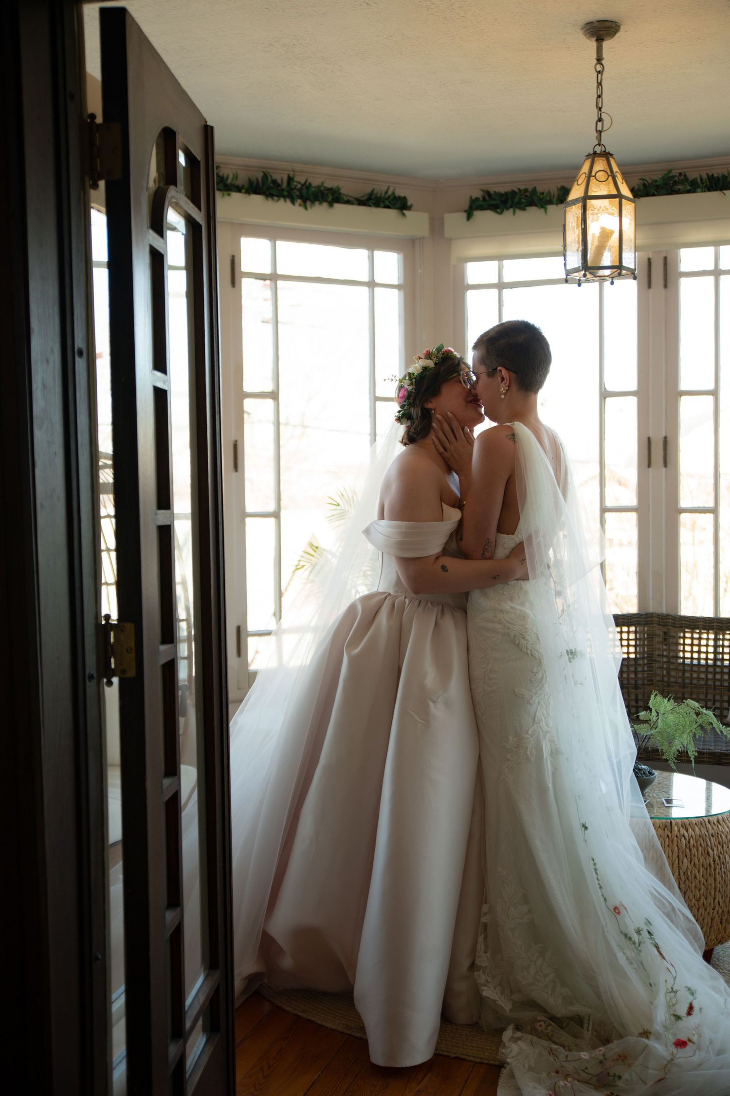 Two brides in white wedding gowns share a tender kiss by sunlit windows adorned with greenery garland.