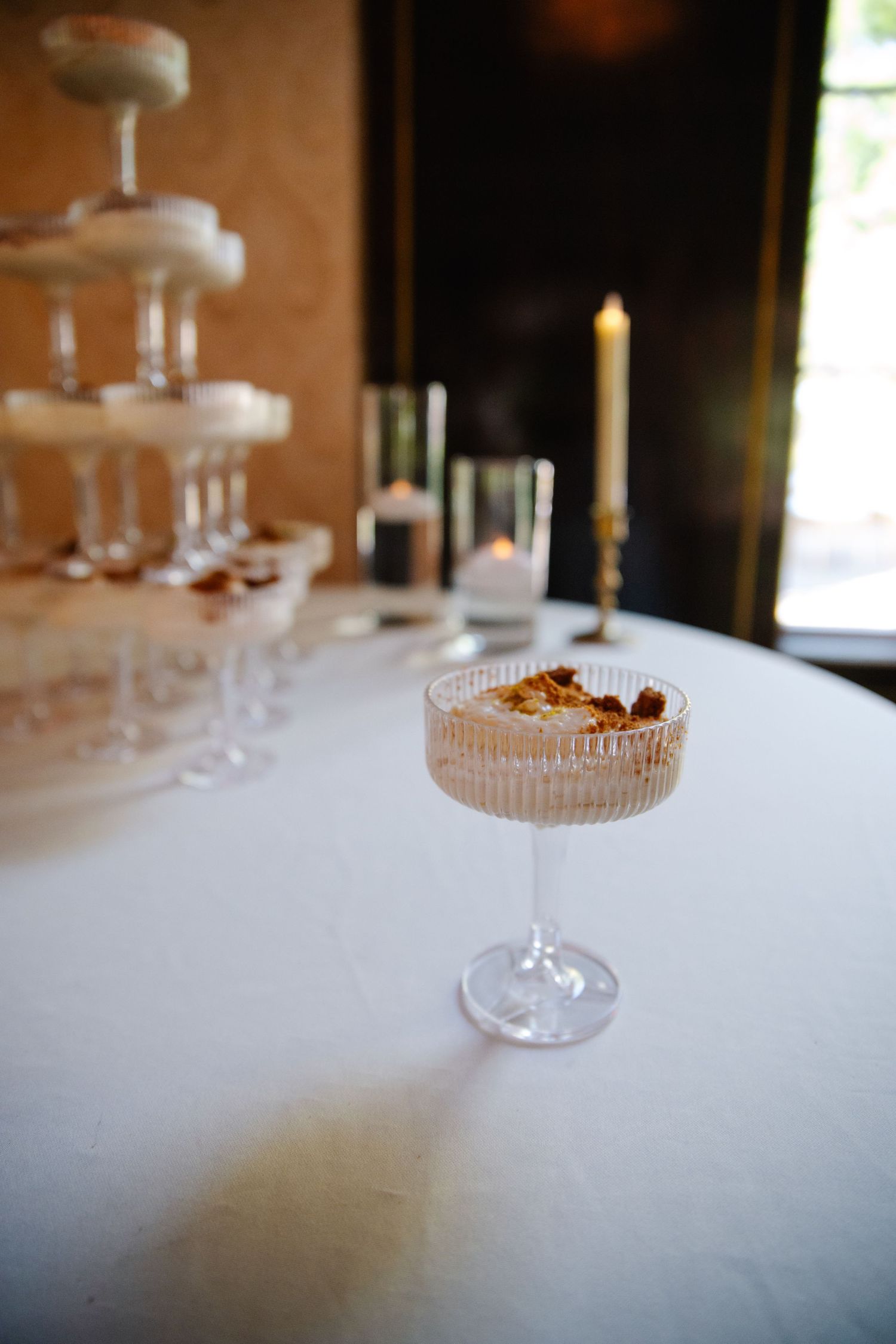 A series of elegant crystal coupe glasses arranged in a row on a white tablecloth in a dimly lit restaurant setting.