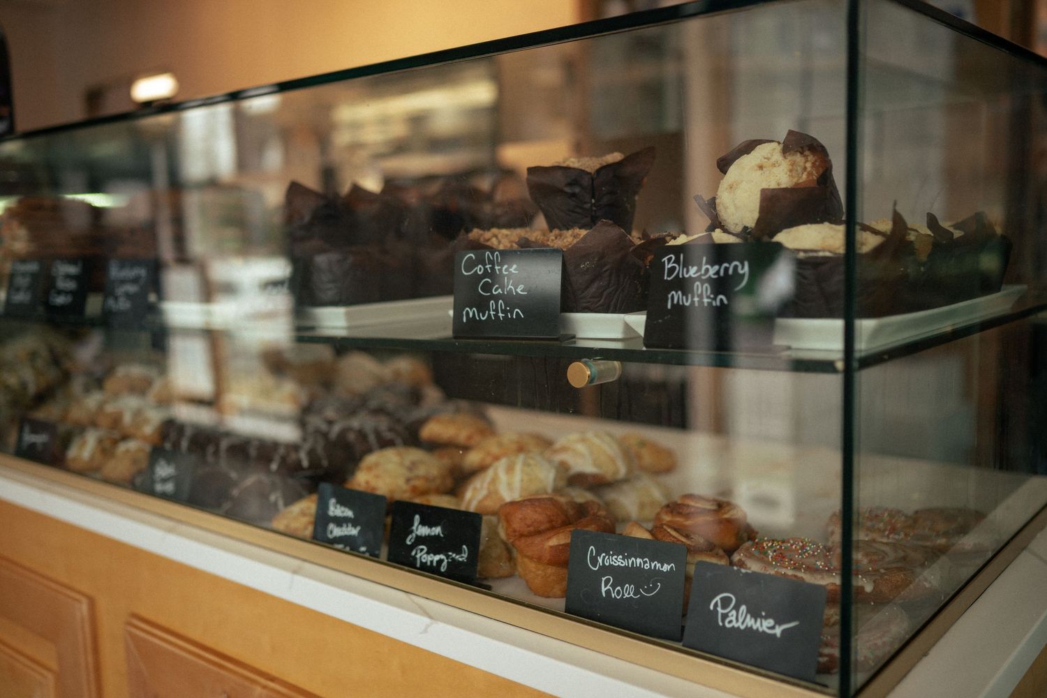 Glass bakery display case filled with muffins, pastries, and baked goods with chalkboard labels.
