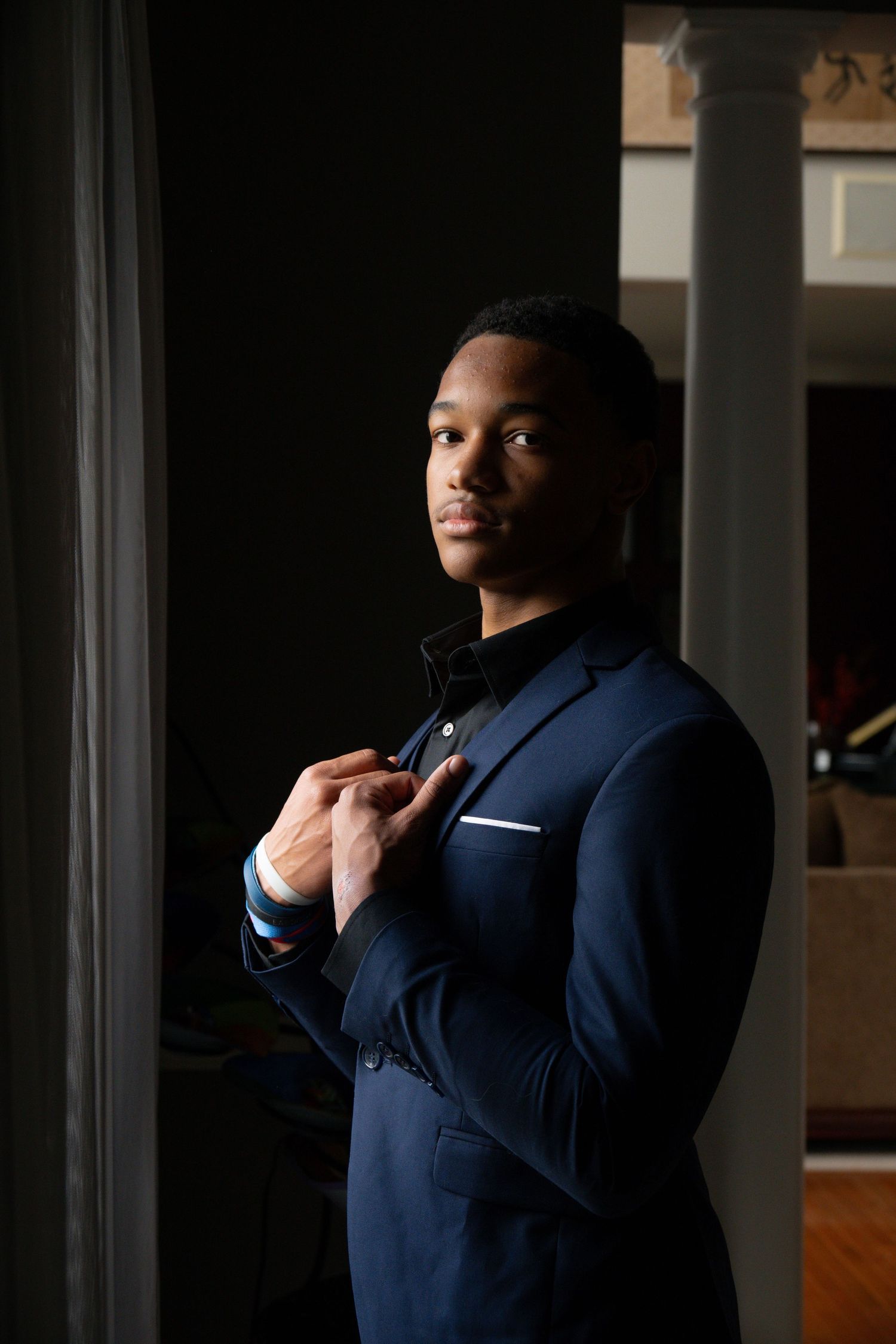 A dramatic portrait shows someone adjusting their navy blue suit tie in window light.