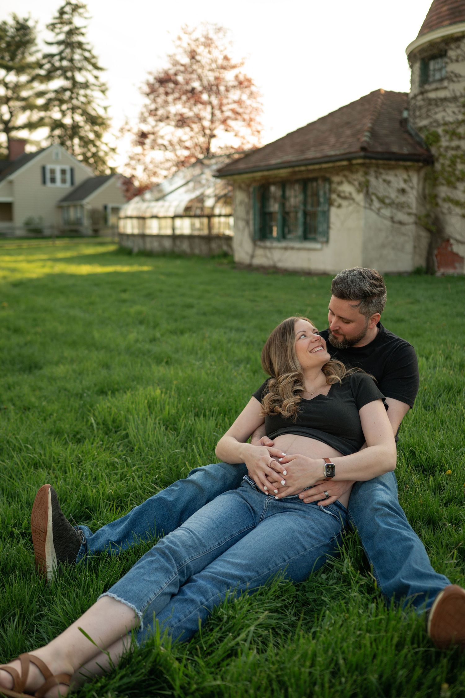 Pregnant couple sitting on green grass, man kissing woman's head while both hold her baby bump, historic building backdrop.